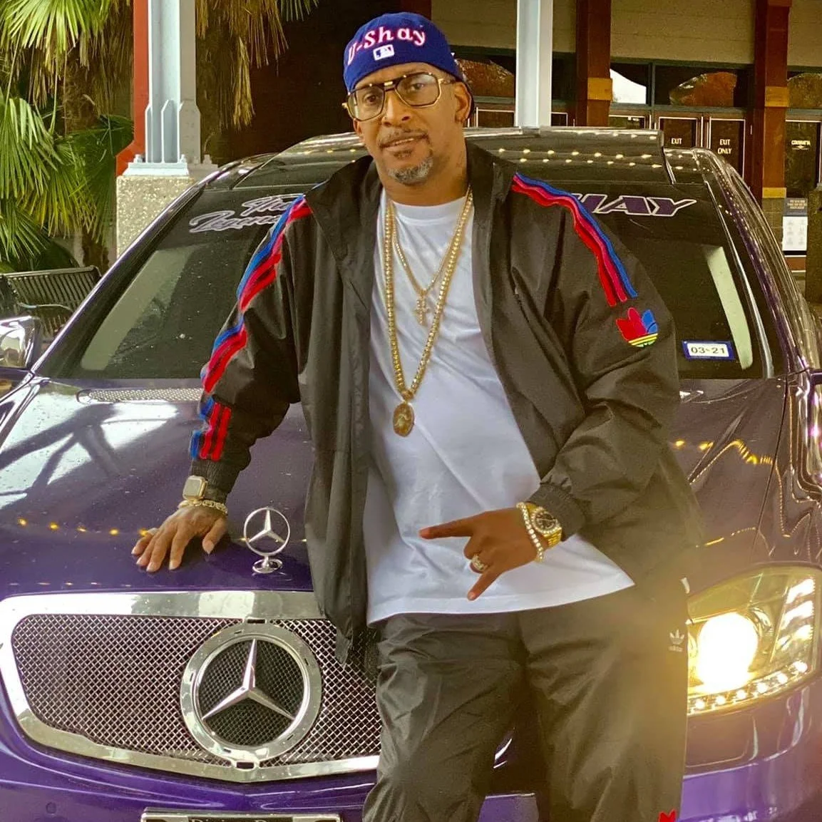 A man dressed in urban streetwear, wearing a blue cap, standing next to a purple Mercedes-Benz car with a large Mercedes emblem on the front grille. The man is wearing glasses, gold chains, a watch, and a black jacket with red and blue stripes and colorful patches. The background features a palm tree and a building with glass doors.