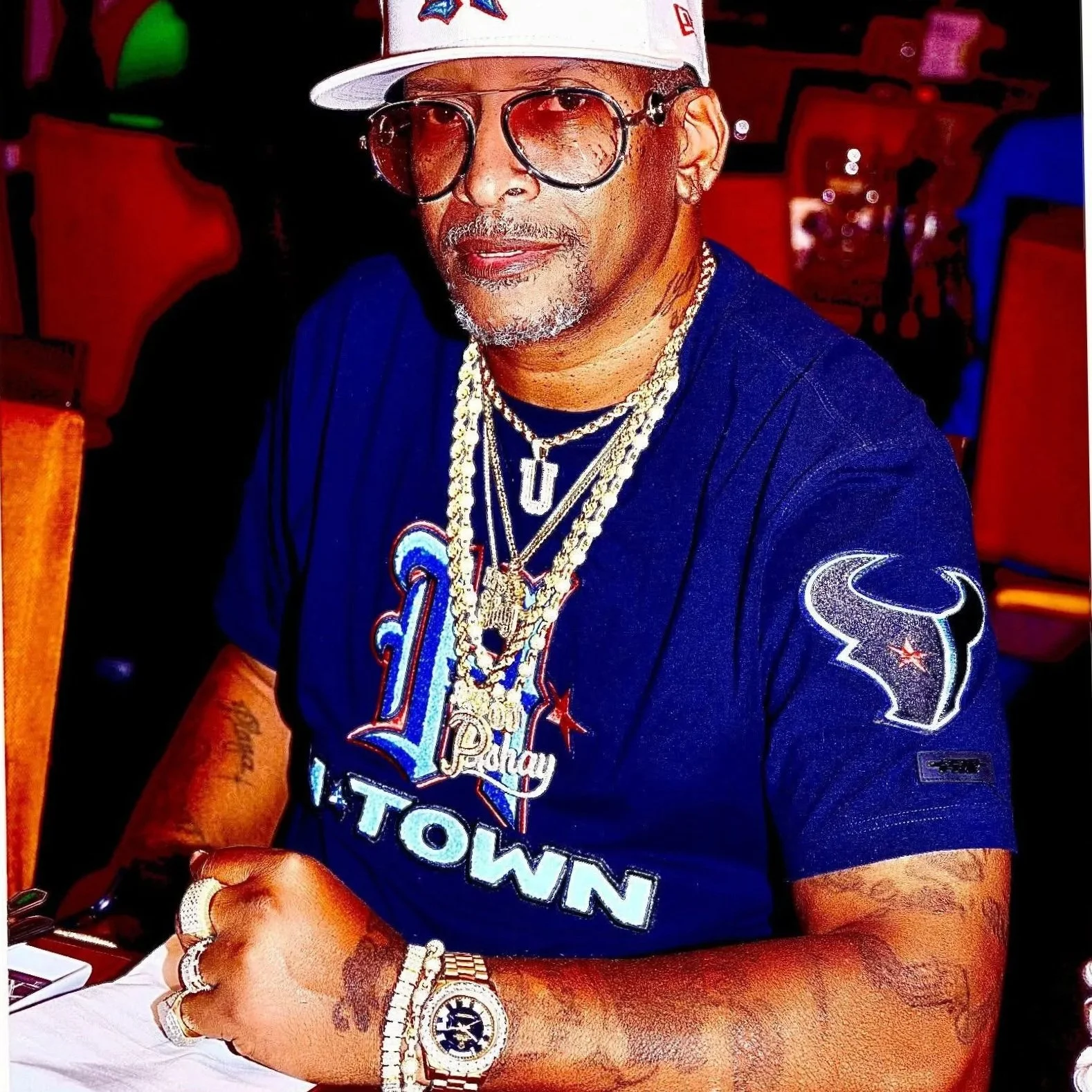 A man with glasses and facial hair wearing a white cap, gold chains, and a blue T-shirt with red and white lettering, sitting at a table with his arms resting on it, in a dimly lit restaurant or club with red and blue chairs in the background.
