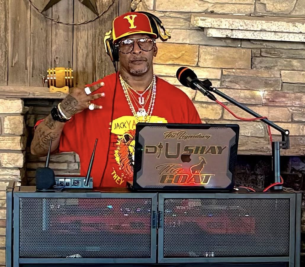 A man dressed in red with a red and yellow baseball cap, multiple necklaces, tattoos, and sunglasses, standing behind DJ equipment, including a laptop with the words "The Legend DJ Shy" and other graphics, in front of a stone fireplace.