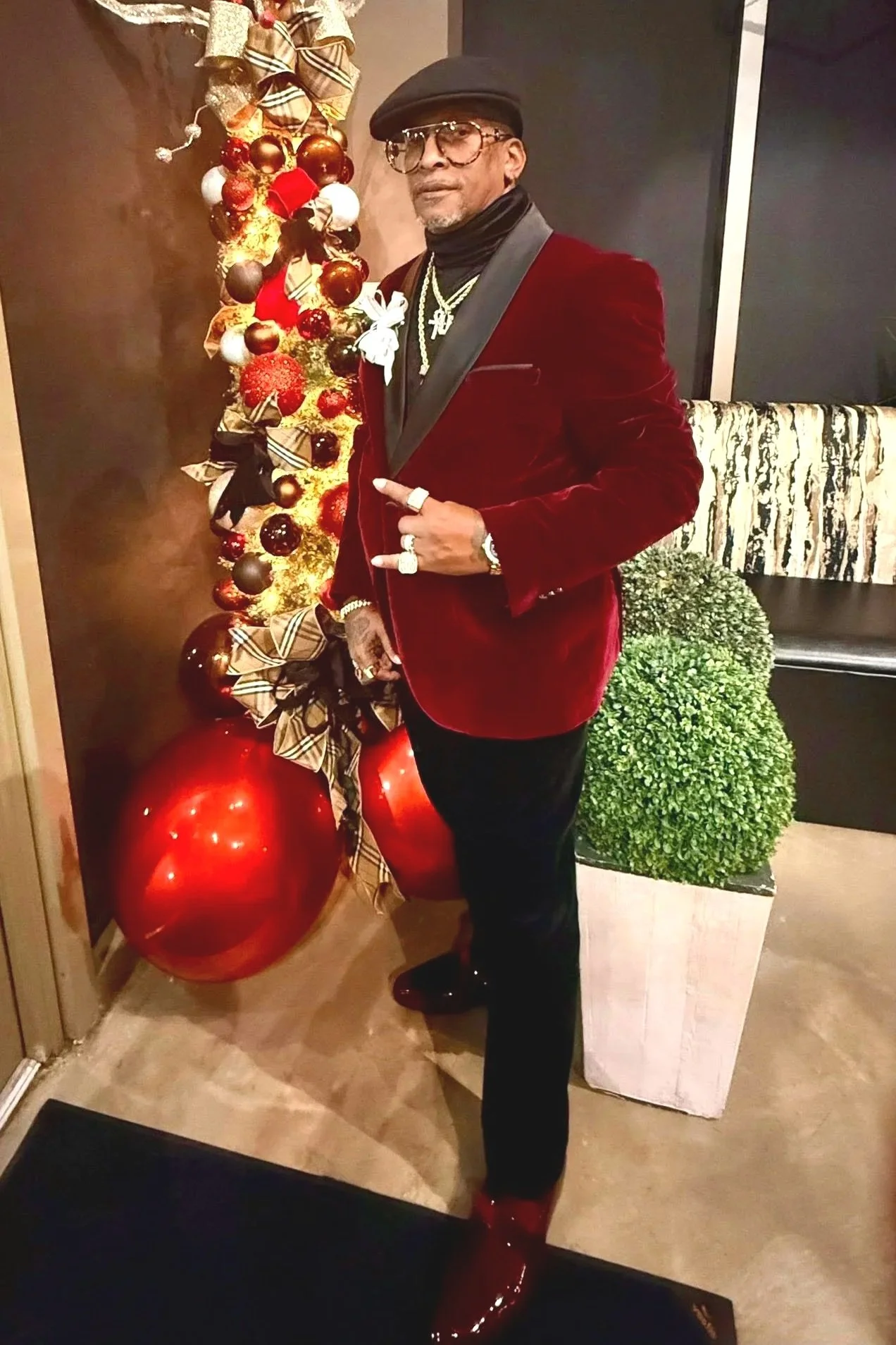 A man in a red velvet blazer, black pants, and red shoes standing next to a decorated Christmas tree with large red ornaments and plaid bow decorations. He is wearing glasses, a black cap, multiple necklaces, rings, and a watch, with a festive posture.