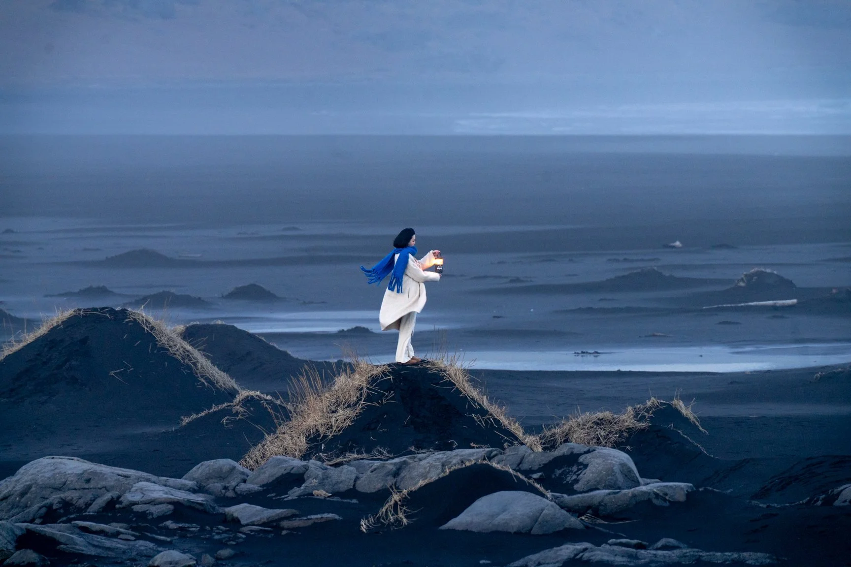 A woman in a white coat and blue scarf holding a lantern stands on a hill overlooking a vast ocean at dusk or dawn, with a cloudy sky and distant waves.