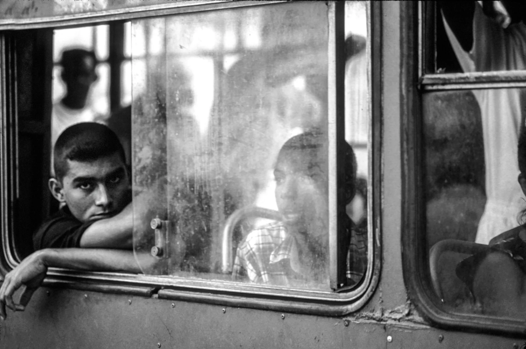 Black and white photo of children sitting inside a bus, seen through a window with a metal frame. One boy with short hair and a serious expression rests his head on his arm, looking out. Other children are seated nearby, some facing the window, some 