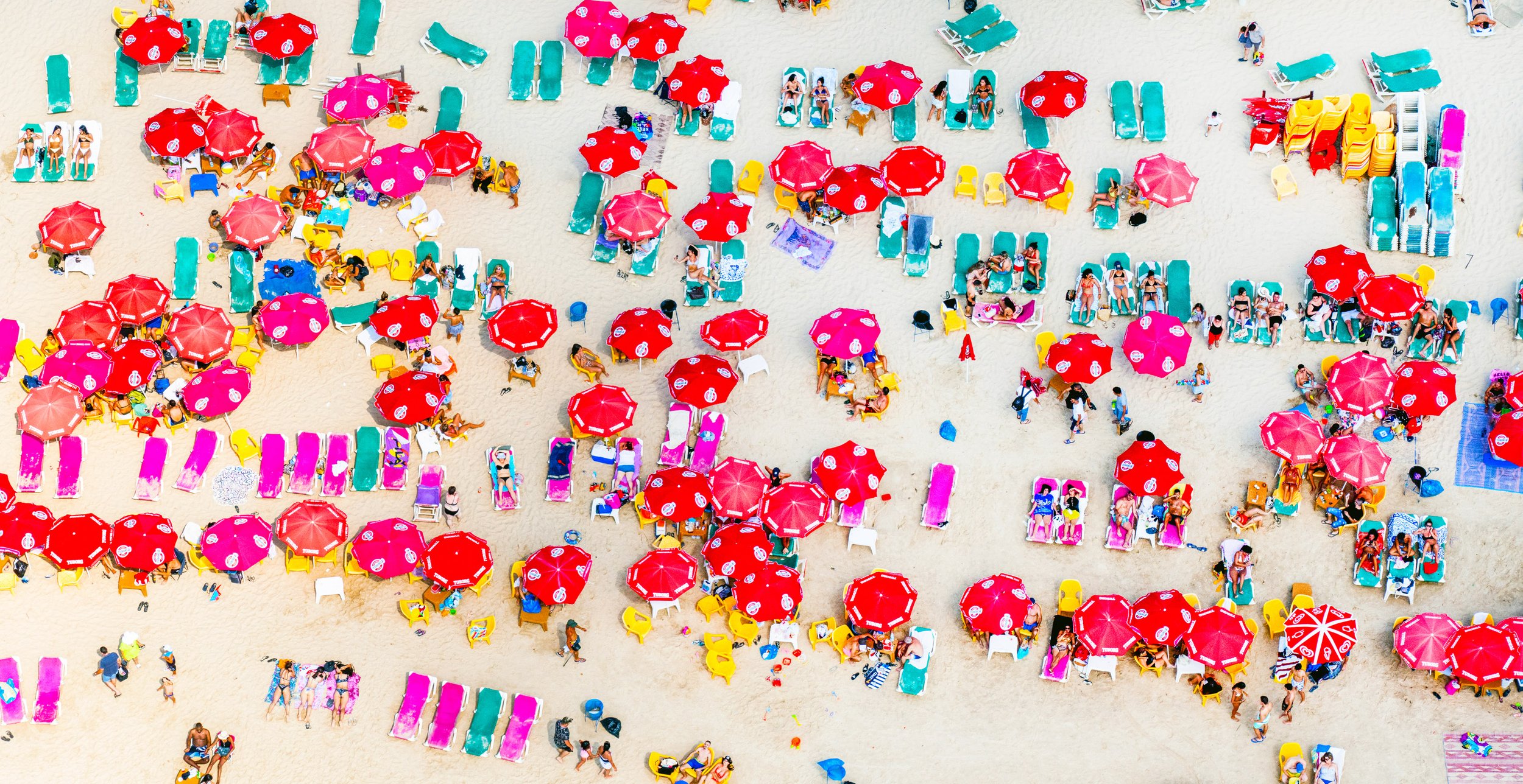 An aerial view of a crowded beach with red and pink umbrellas, sunbathers on lounge chairs, and groups of people relaxing on the sand.