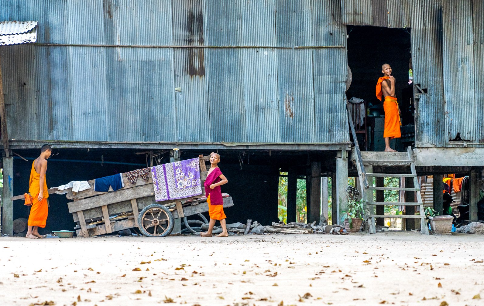 Three children in orange traditional clothing playing outside a stilted house made of corrugated metal sheets.