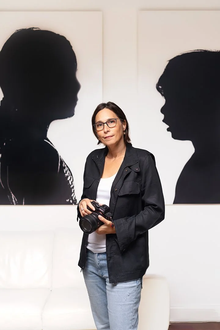Woman standing in front of two large black silhouettes of children on white background, holding a camera, wearing glasses, black jacket, white shirt, and jeans.