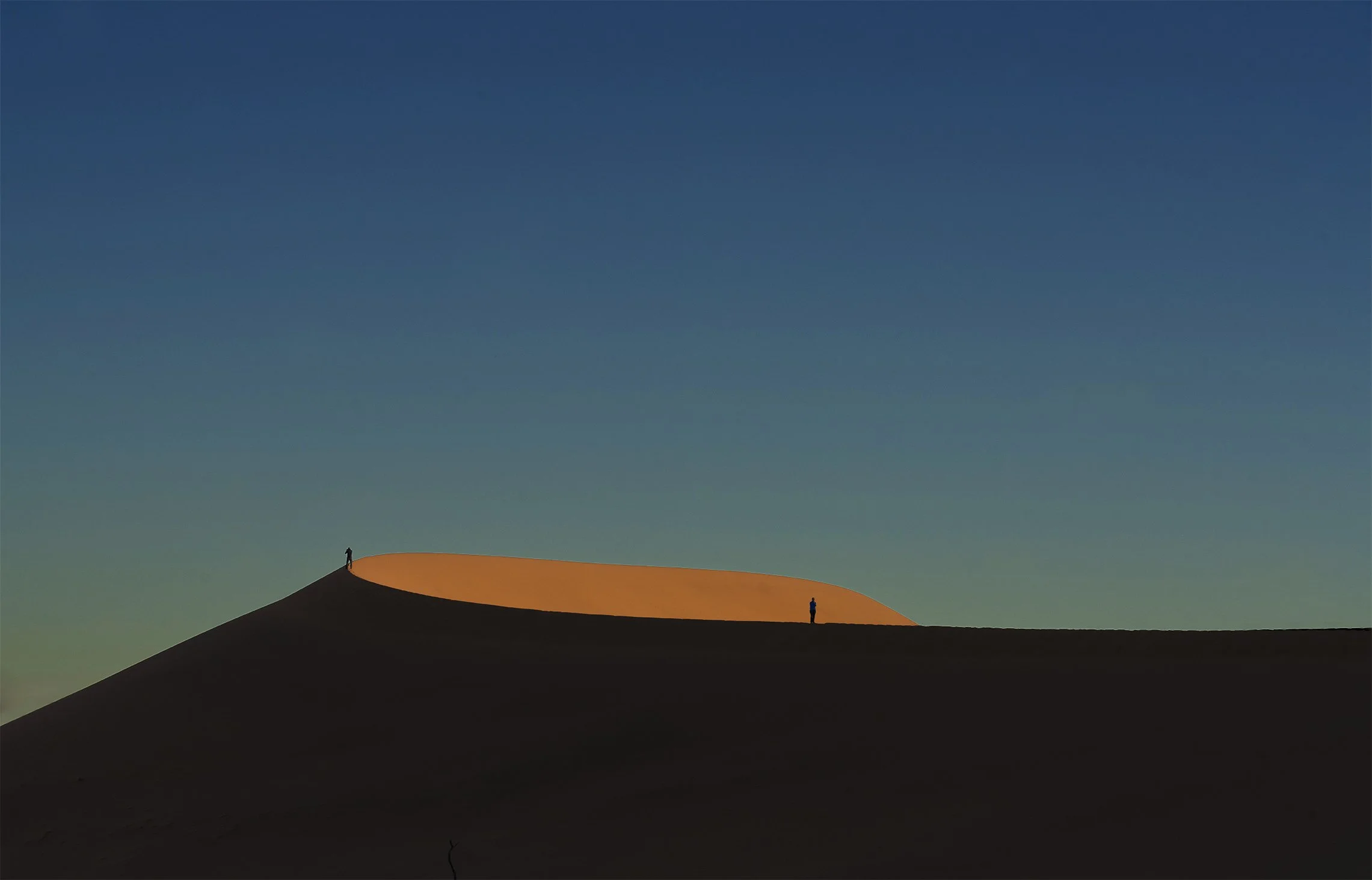 Silhouettes of two people standing on a large, curved sand dune at sunset or sunrise, with a gradient sky transitioning from deep blue to light green.
