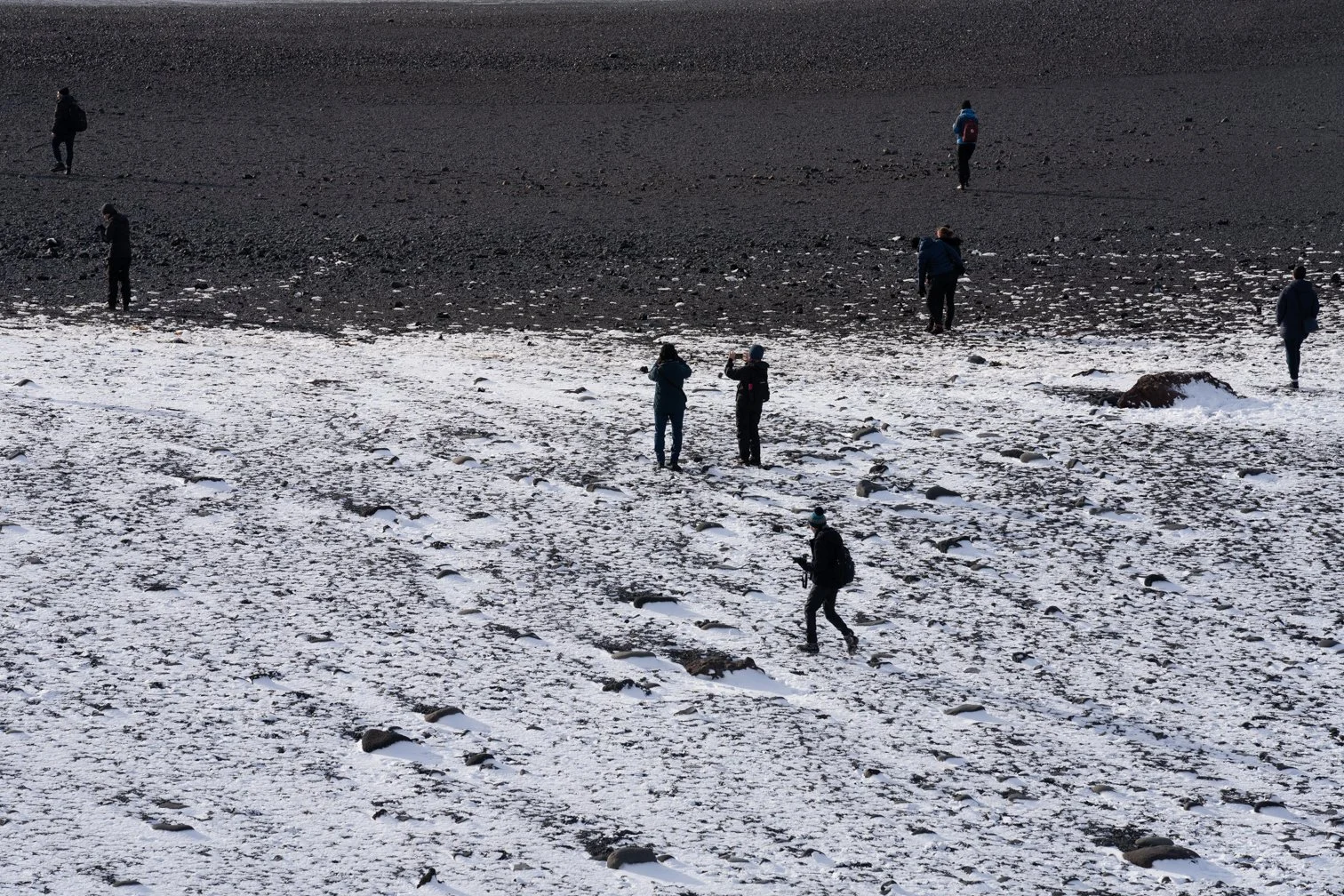Multiple people walking on a snow-covered landscape with patches of dark ground visible in the background.