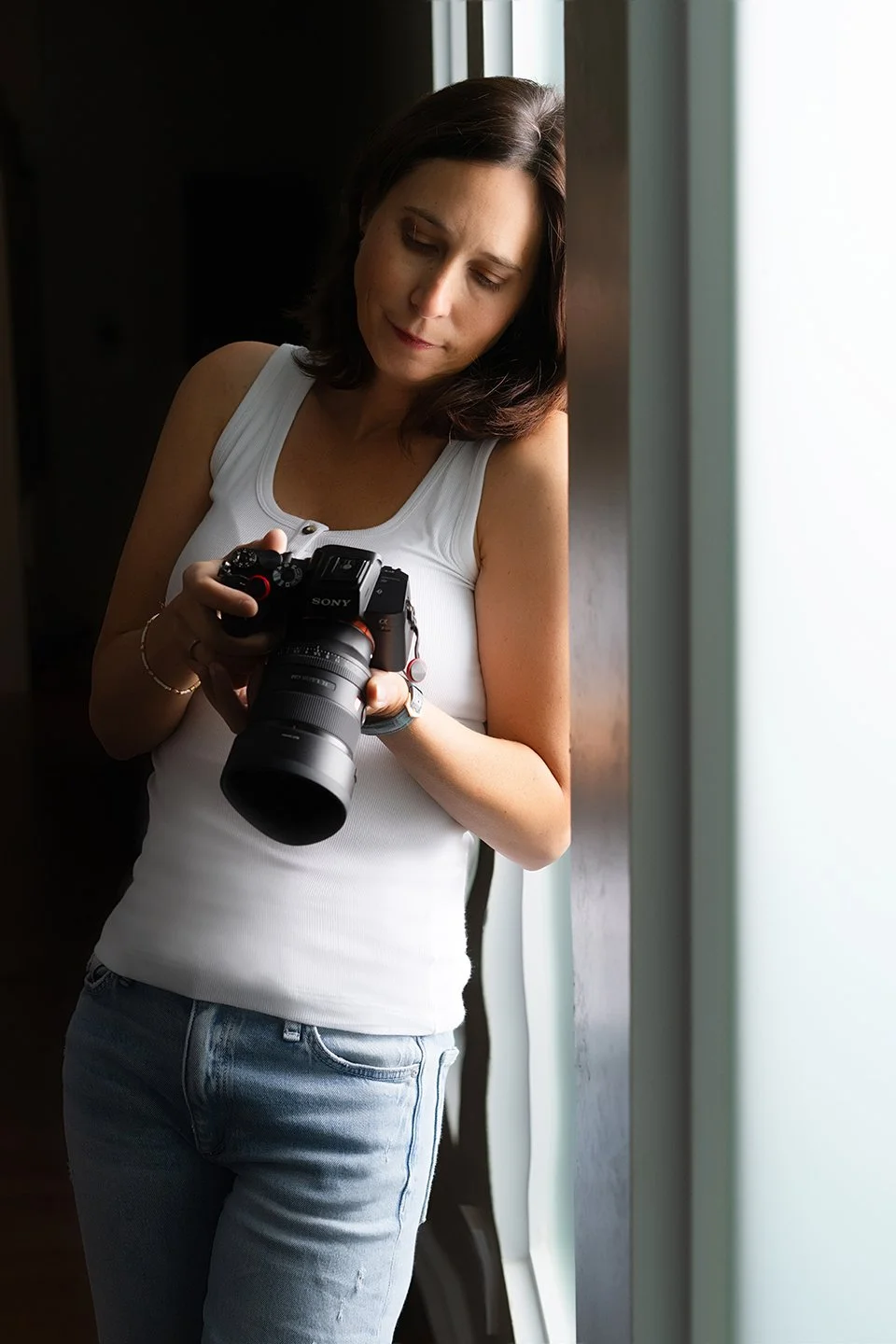 A woman with dark hair looking down at a camera she is holding near a bright window.