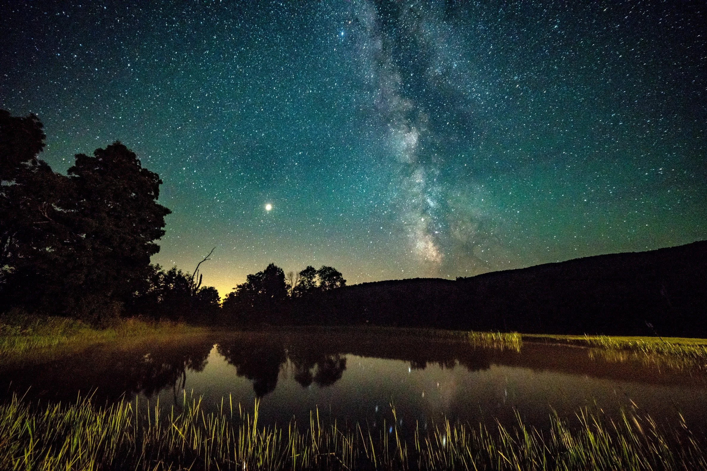 Night sky filled with stars and the Milky Way galaxy, with silhouettes of trees and a reflection of the sky on a body of water in the foreground.