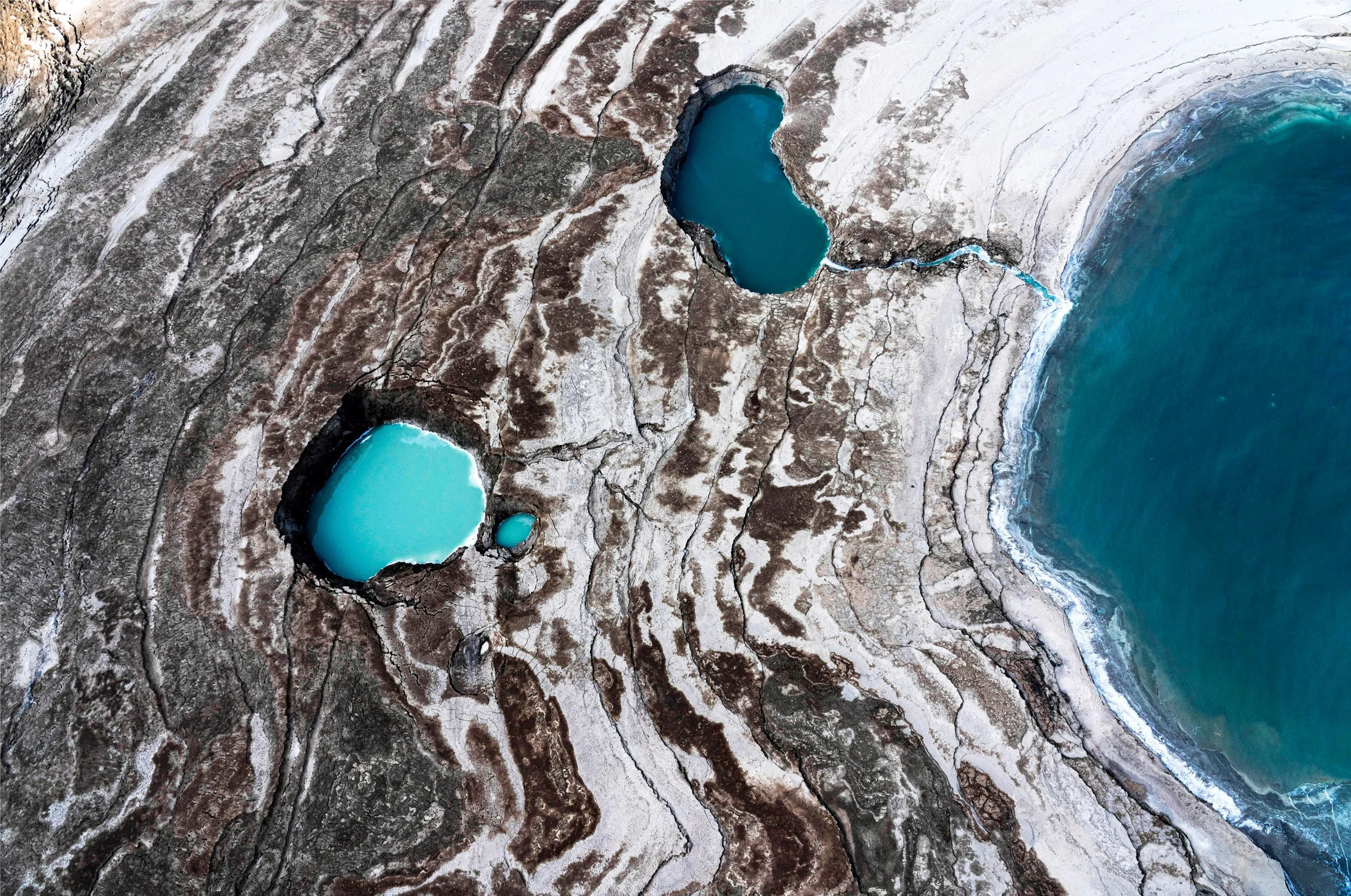 Aerial view of a landscape with two blue lakes, one larger and one smaller, surrounded by land with rocky and cracked terrain, next to a body of water on the right side.