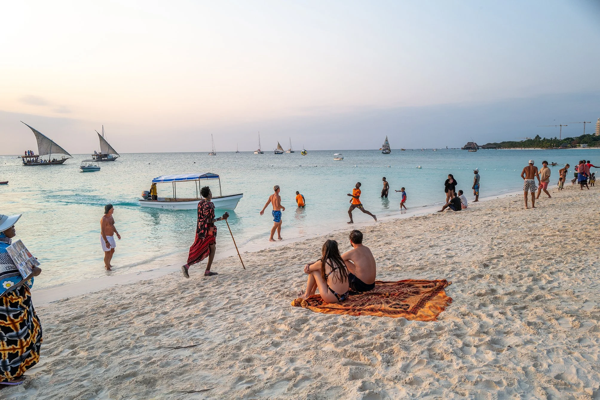 A busy beach scene at sunset with people relaxing on the sand, children playing, and boats sailing on the calm water. There are sailboats and paddleboats in the distance, and some individuals walking along the shoreline.