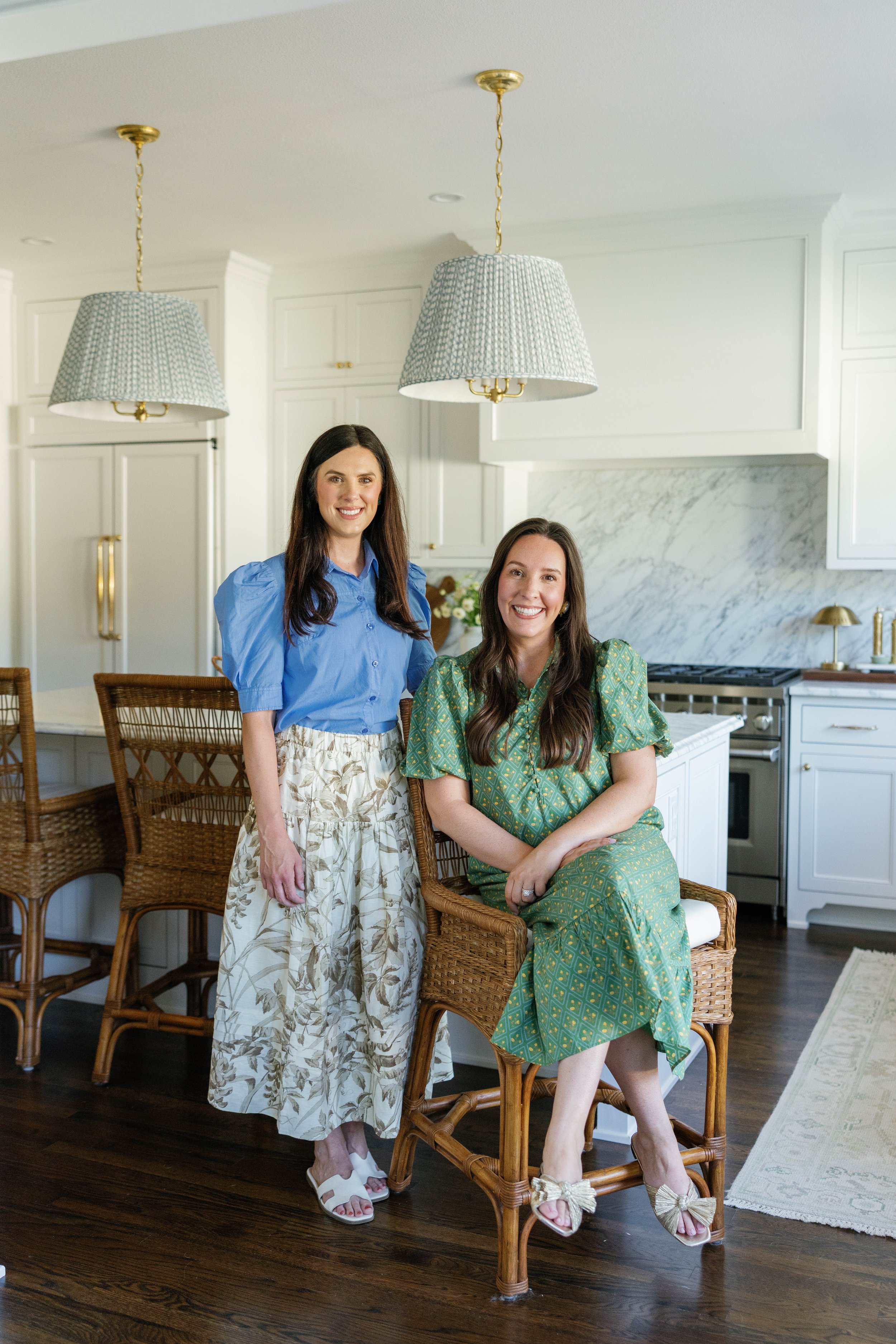 Two women in colorful dresses pose in a bright kitchen with two large pendant lights and wooden chairs, one sitting on a chair and the other standing behind her.