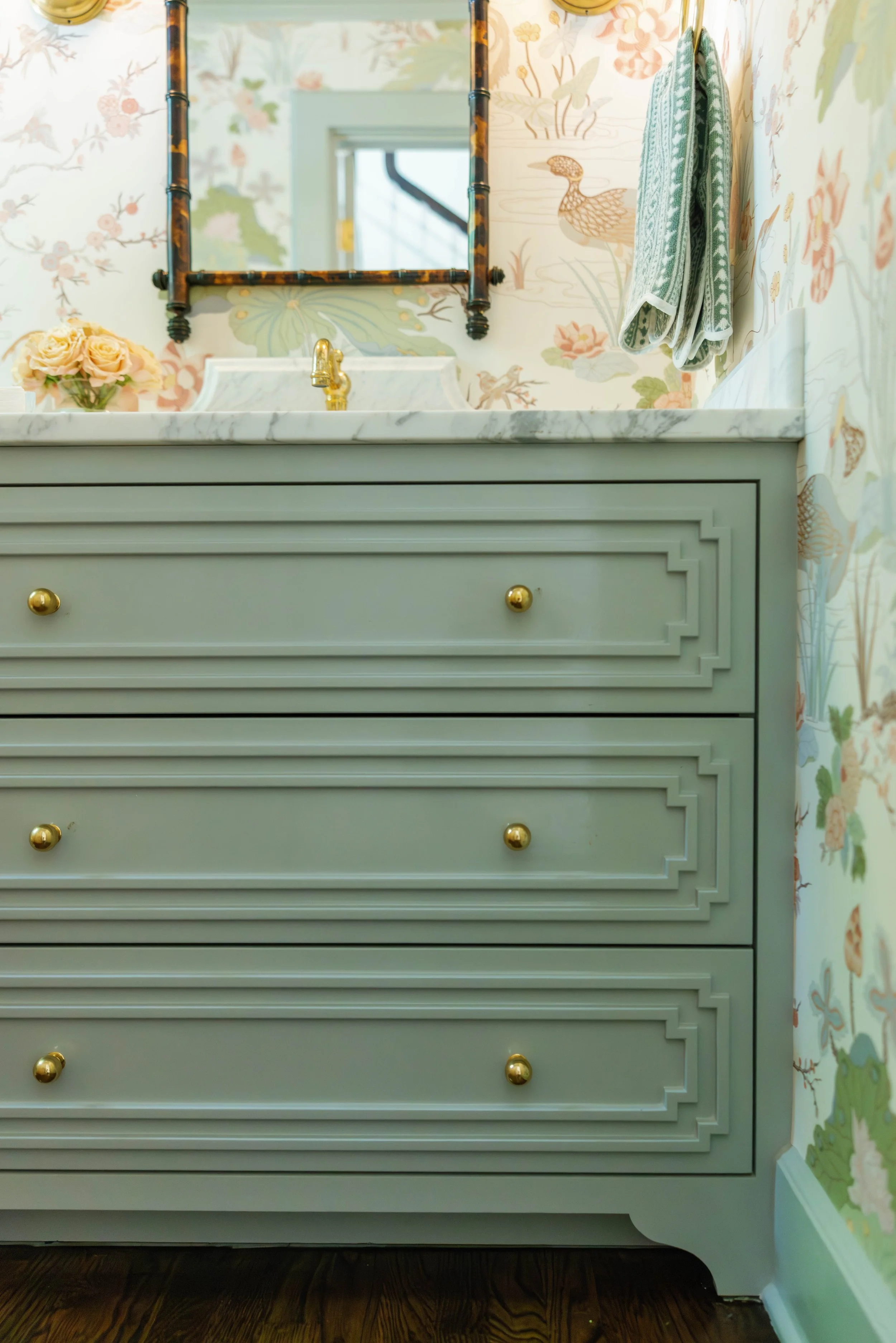 Close-up of a bathroom vanity with a marble countertop, gold faucet, light gray cabinet drawers with gold knobs, floral wallpaper, a mirror with a tortoise shell frame, a small bouquet of roses, and towels hanging on the wall.