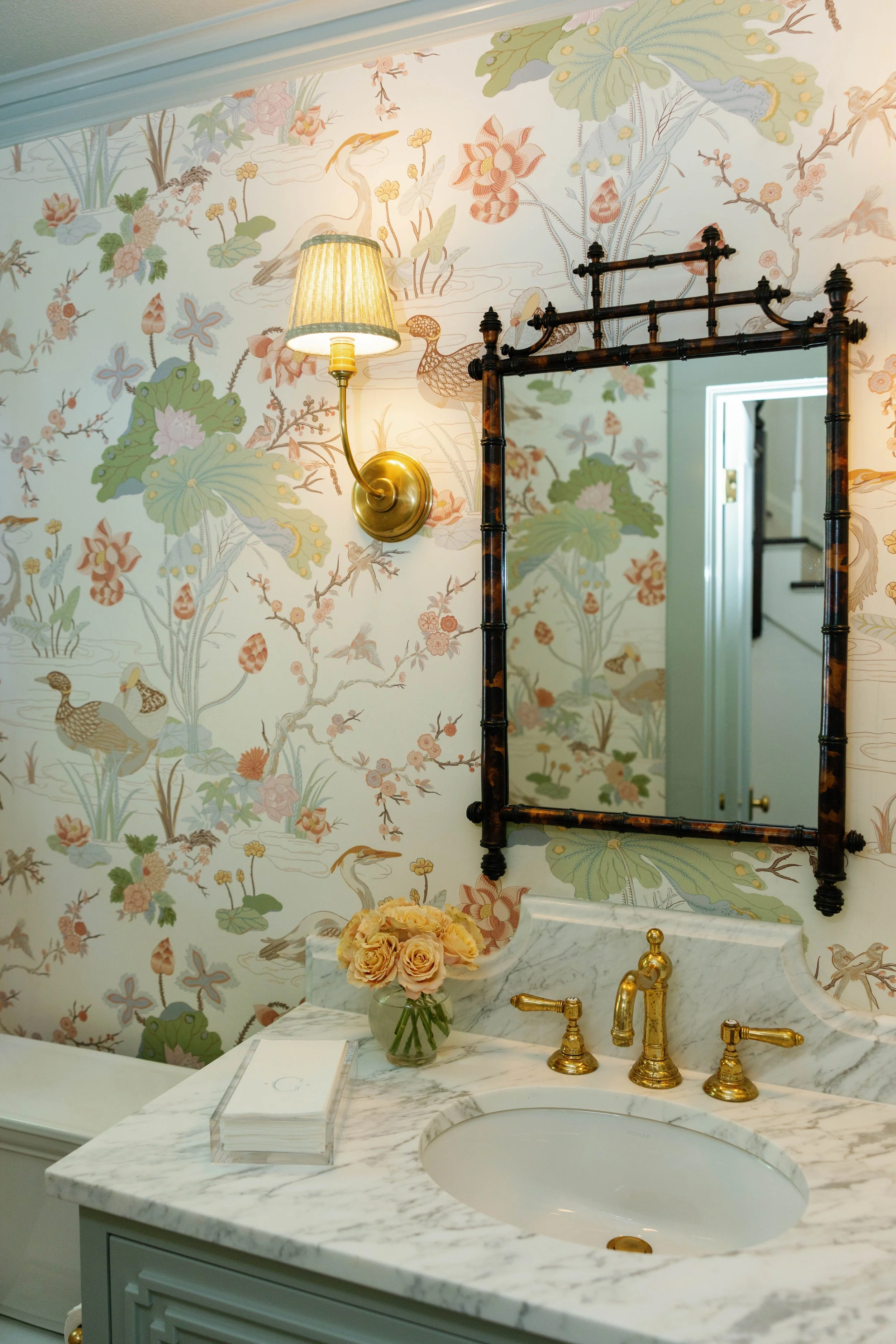 A bathroom vanity with a marble countertop, gold fixtures, and a floral wallpaper background featuring cranes, lotus leaves, and flowers. There is a mirror frame, a lamp, a vase with peach roses, and a white box on the counter.