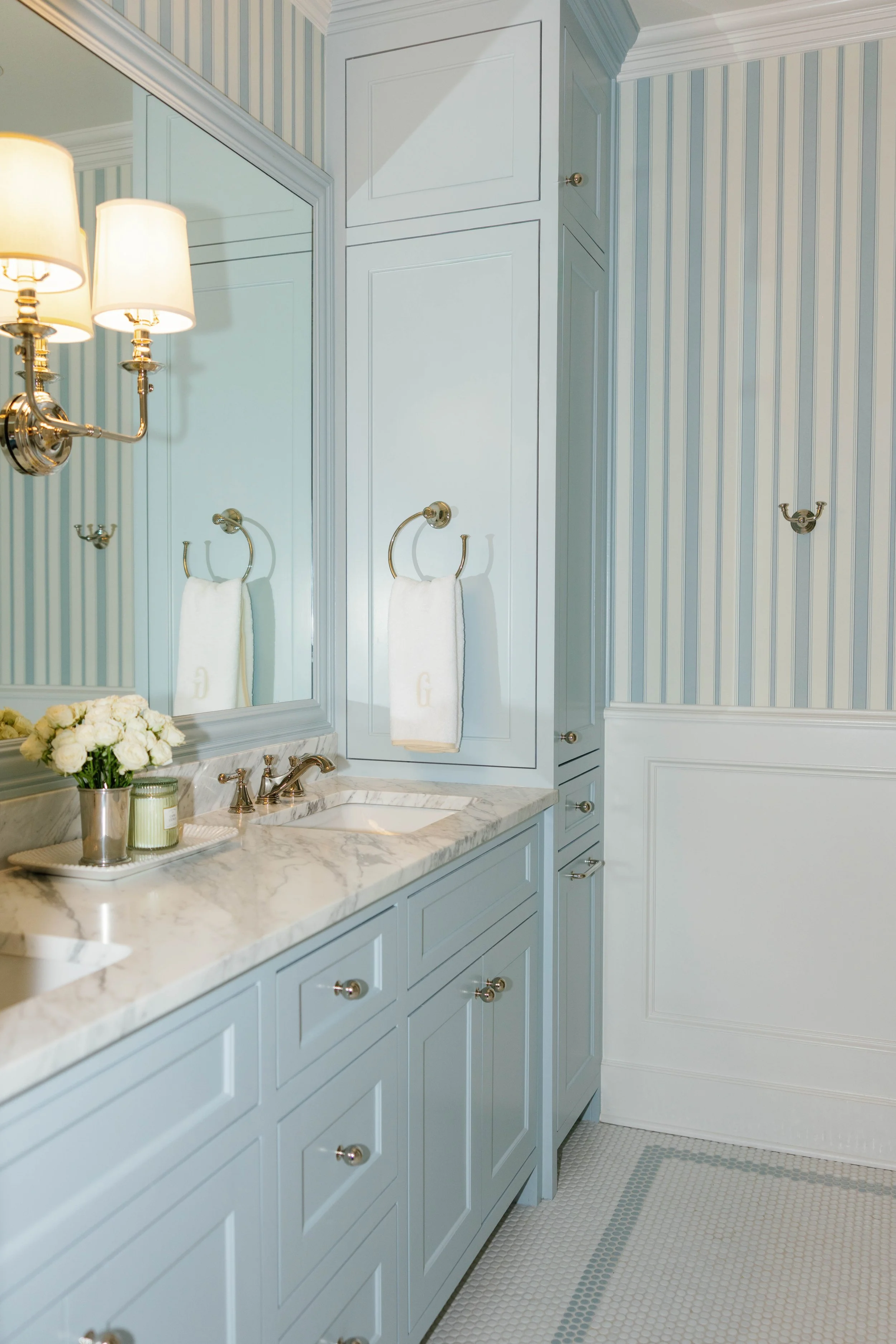 Blurred view of a light blue bathroom with cabinetry, a marble countertop, wall-mounted towel rings with white towels, a mirror, wall-mounted light fixtures, a small bouquet of white flowers, and a patterned tile floor.