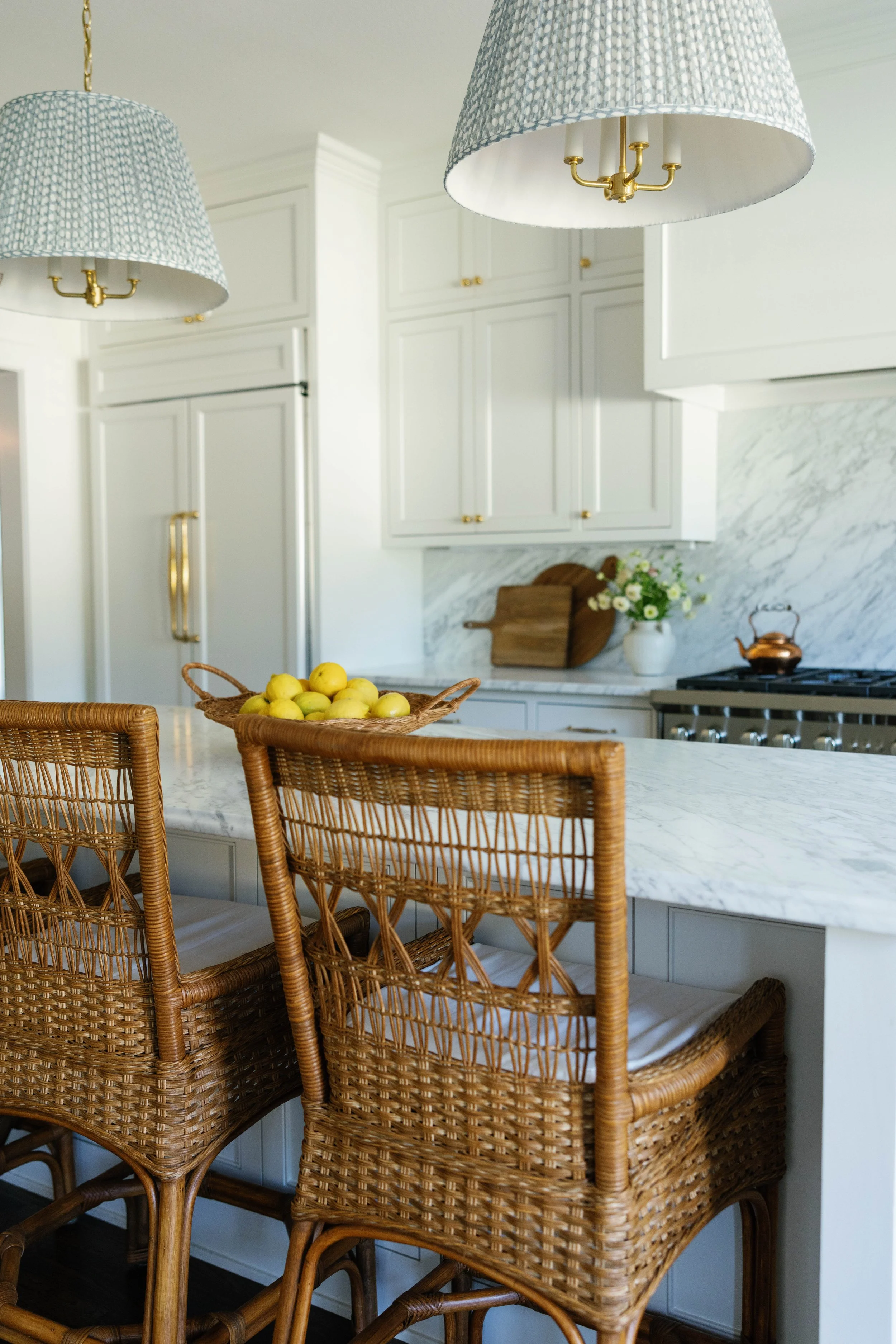 Kitchen with white cabinets, marble countertop, and woven rattan chairs around a marble island. Two hanging light fixtures with blue patterned shades, a cutting board, and a vase of flowers in the background.