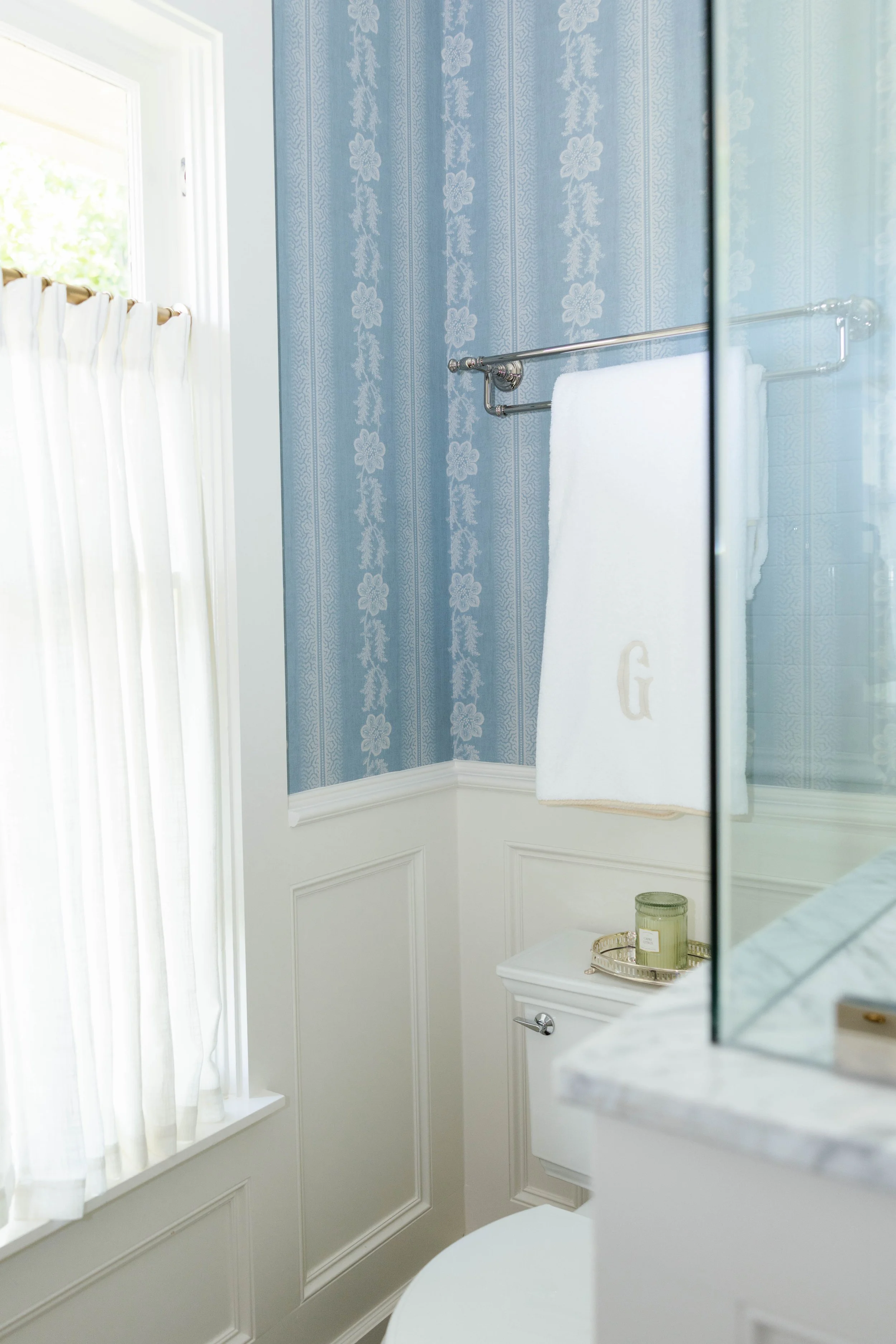 A bathroom with light blue floral patterned wallpaper, a white window with sheer curtains, a silver towel bar with a white towel, a small white sink, a round mirror, and decorative items like candles on the countertop.