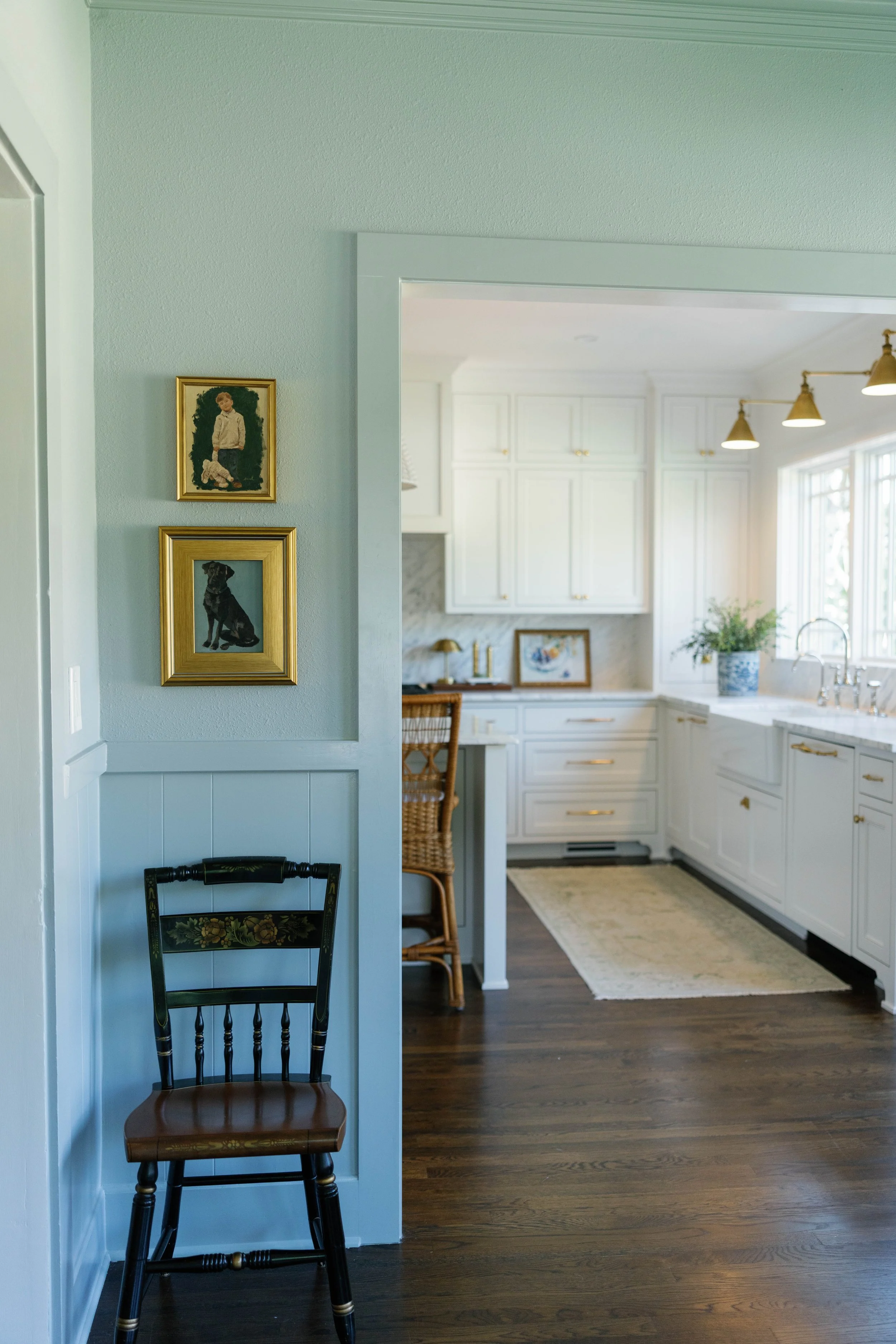Interior view of a bright, white kitchen with a plant on the counter, floral rug, and dark wood flooring. A framed picture of a dog and a child is on a wall.