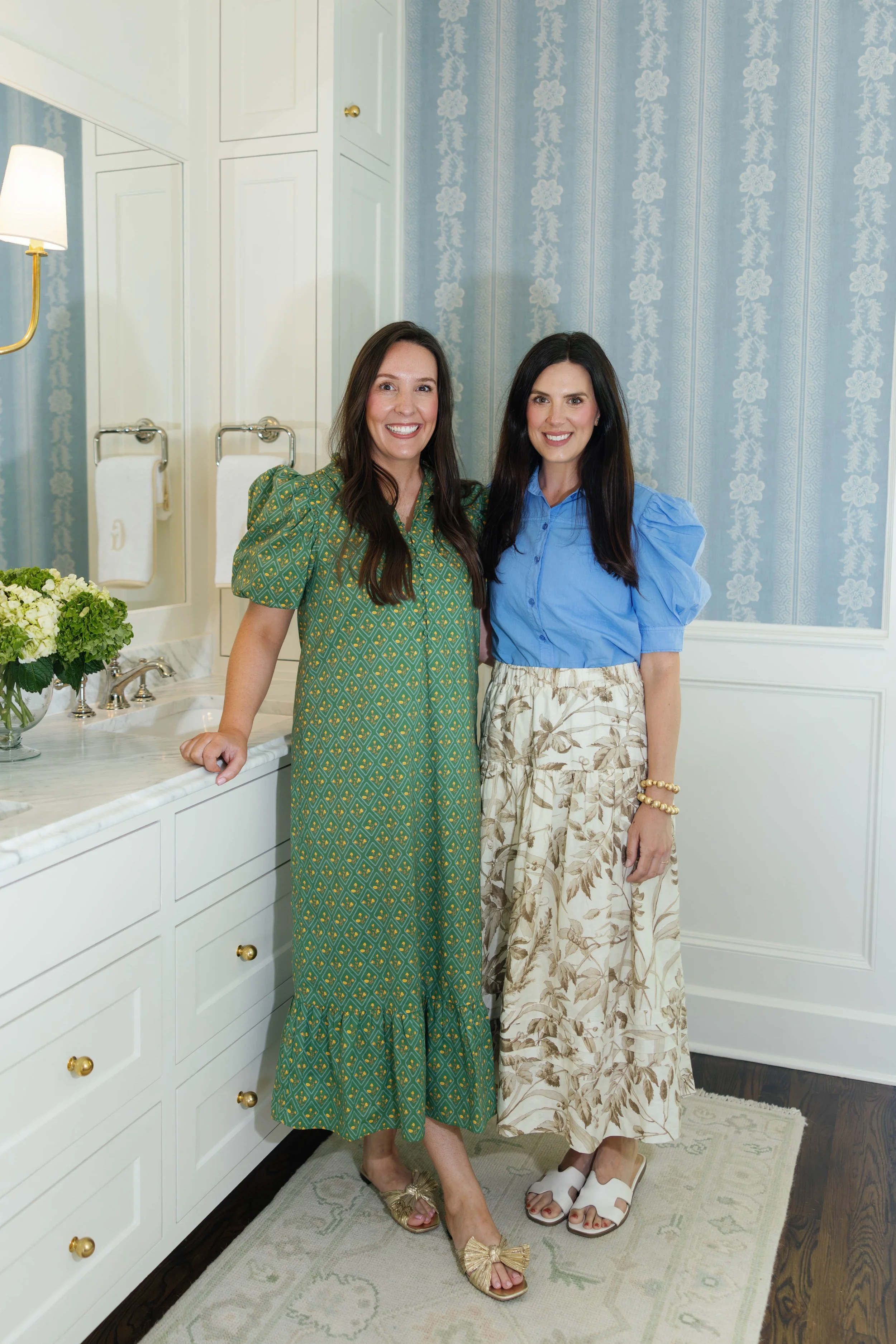 Two women standing inside a room with light blue patterned wallpaper, white cabinetry, and a white countertop with flowers. The woman on the left is wearing a green patterned dress and gold sandals with bows, while the woman on the right is wearing a blue top and a beige floral skirt with white sandals. They are smiling and posing for the photo.