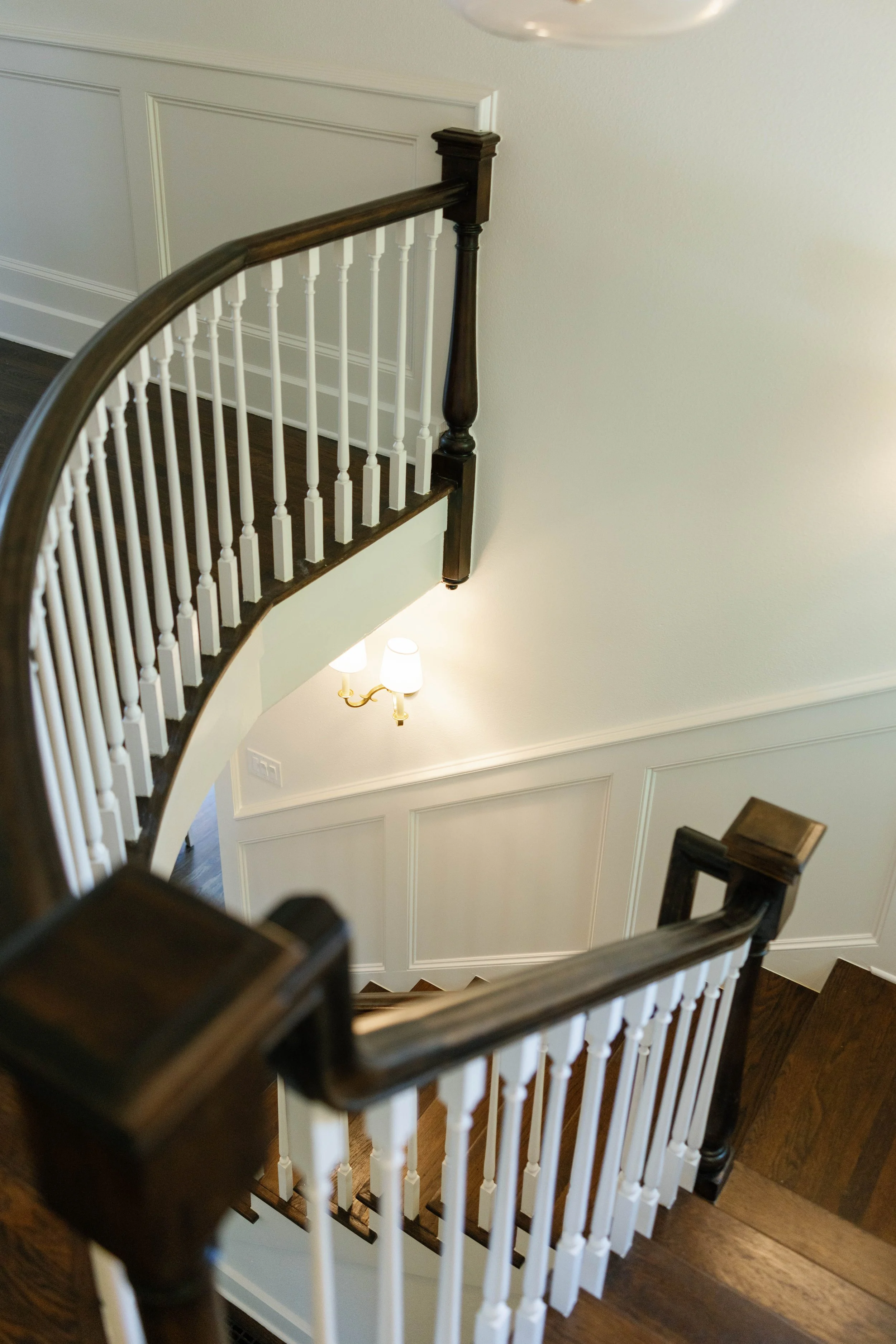 Interior view of a staircase with white railings, dark wooden handrails and posts, and a wall with decorative paneling and a wall-mounted light fixture.