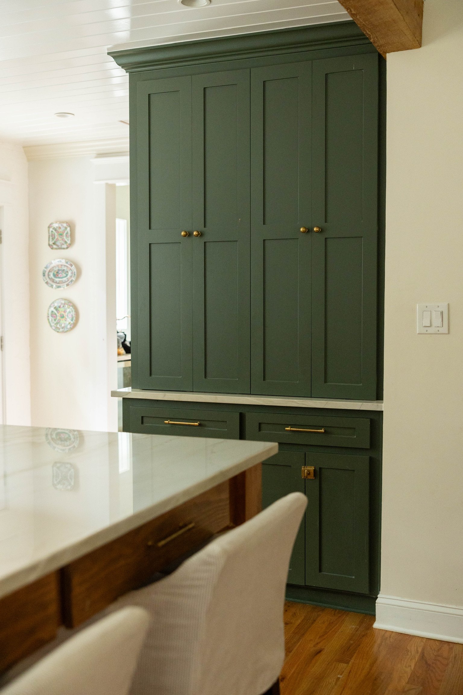 A view of a kitchen with a green cabinet and a wooden counter with a white marble top. The cabinet has brass knobs and is placed against a white wall with a light switch. Part of a chair and a kitchen counter are visible in the foreground.