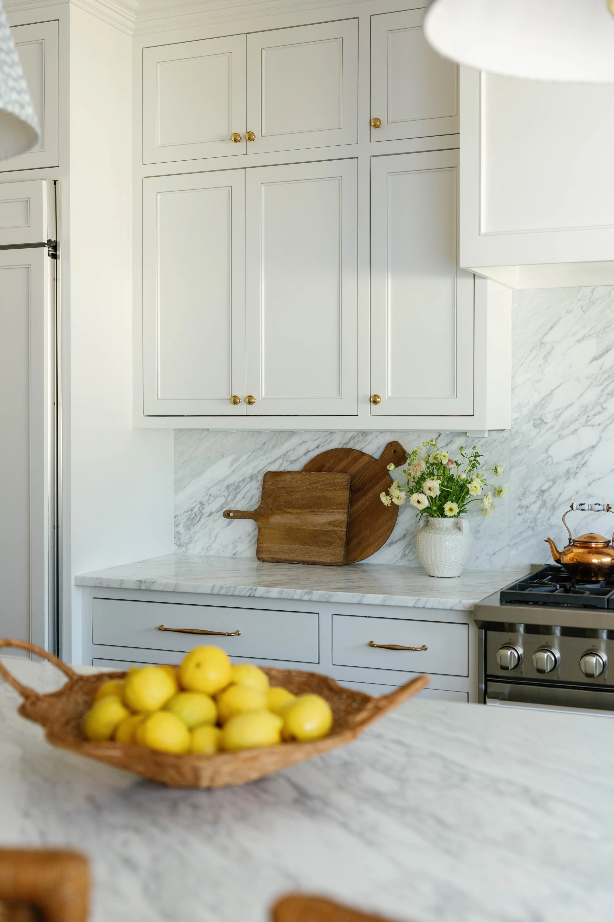 A white kitchen with wood accents, including white cabinets with gold knobs, a marble countertop and backsplash, a wooden cutting board, a white vase with flowers, and a stainless steel stove. The foreground features a basket of yellow lemons.