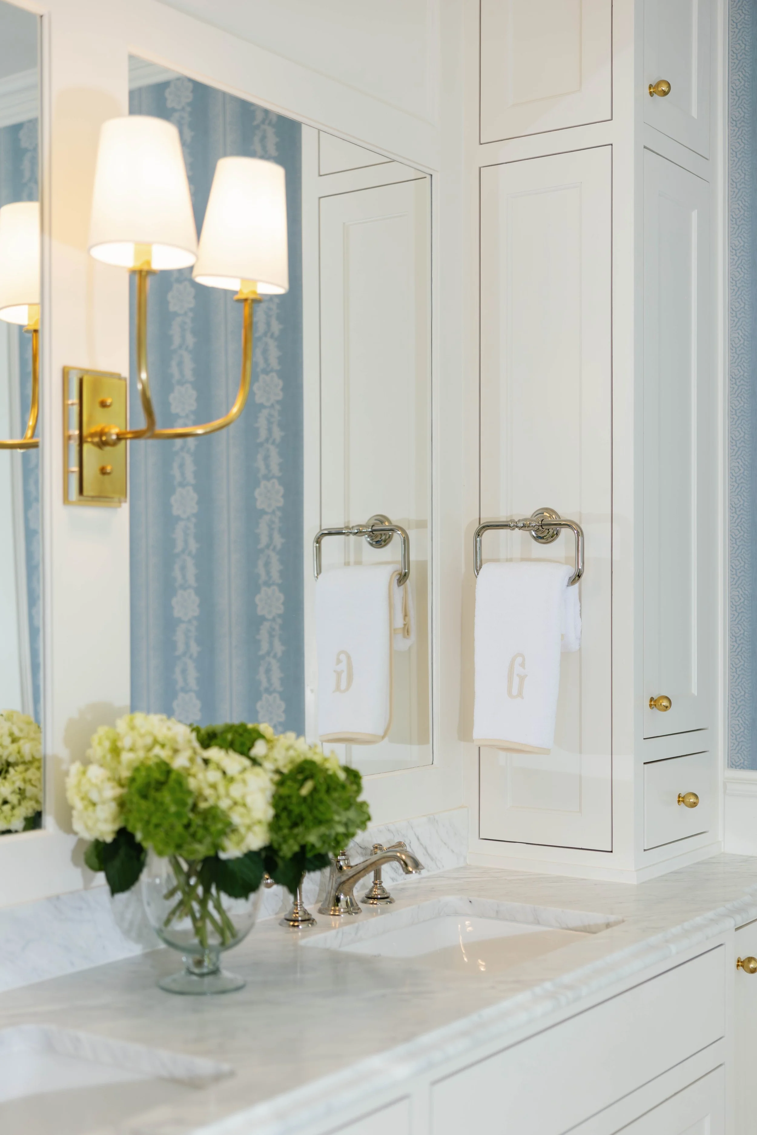 Close-up of a white marble bathroom vanity with a large mirror, a vase of white and green hydrangeas, and a gold wall sconce with three lamps. A towel with monogrammed initials hangs on a towel bar.