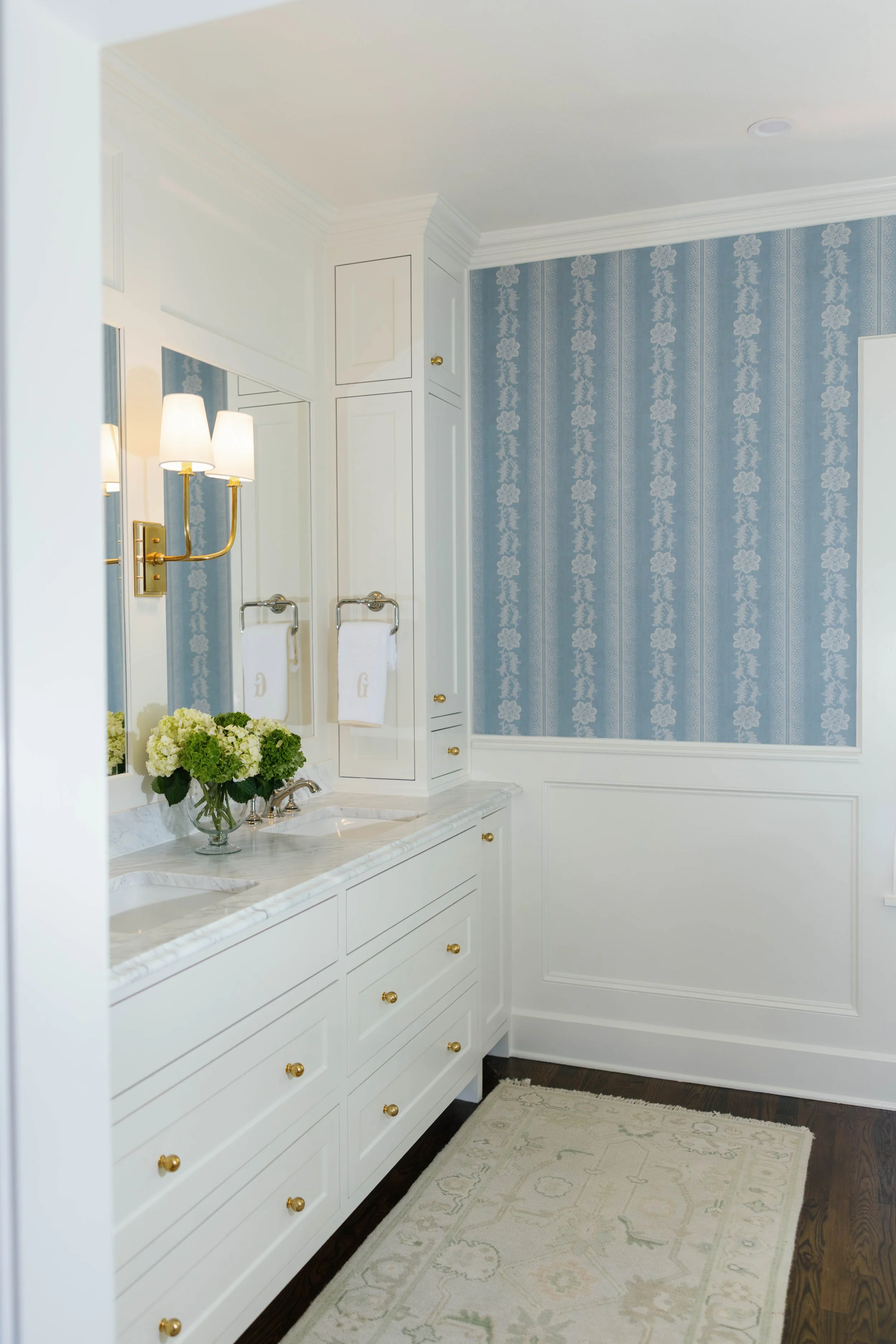 A bathroom with blue patterned wallpaper, white cabinetry with gold knobs, a marble countertop, a mirror, and a wall-mounted light fixture with three lampshades. There is a white towel hanging on a gold towel bar, a vase with green and white flowers on the counter, and a beige patterned rug on dark wooden floor.
