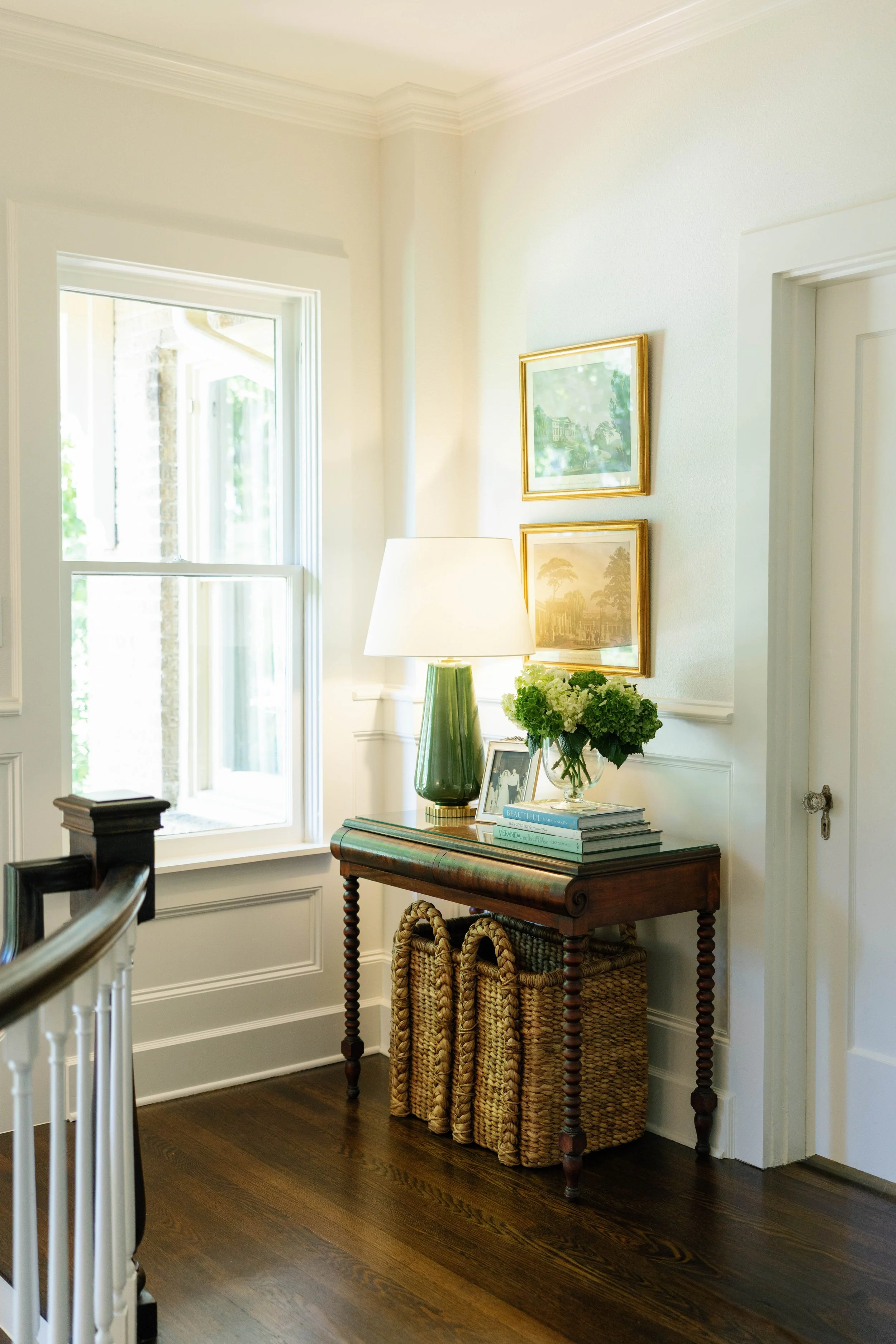 Interior of a bright, elegant living room with a window, table with books, a green lamp, framed artwork, and a vase of white hydrangeas on a wooden table.