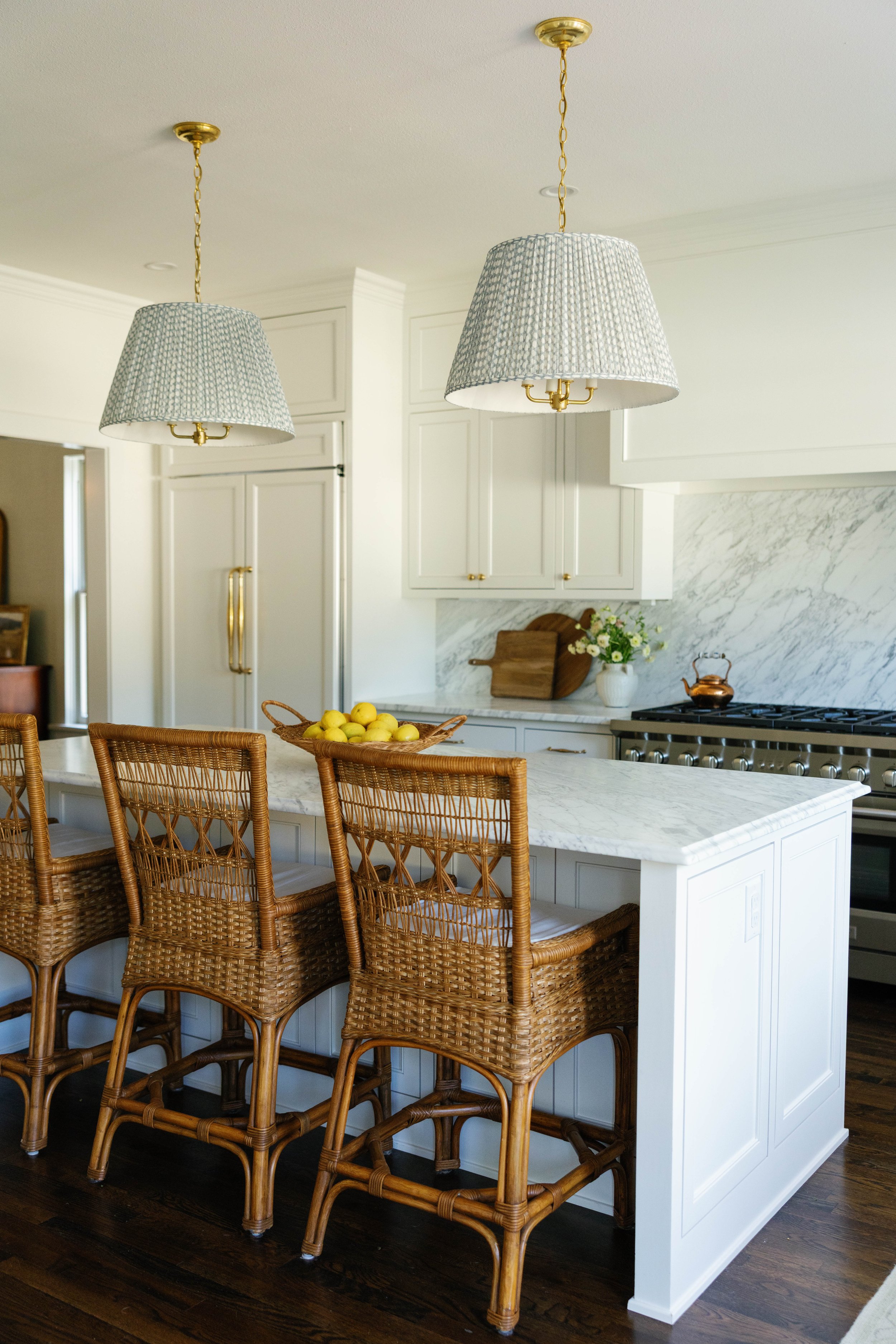 A kitchen with white cabinets, a marble backsplash, and a white island with a basket of lemons. Woven rattan chairs are around the island, and two pendant lights hang from the ceiling.