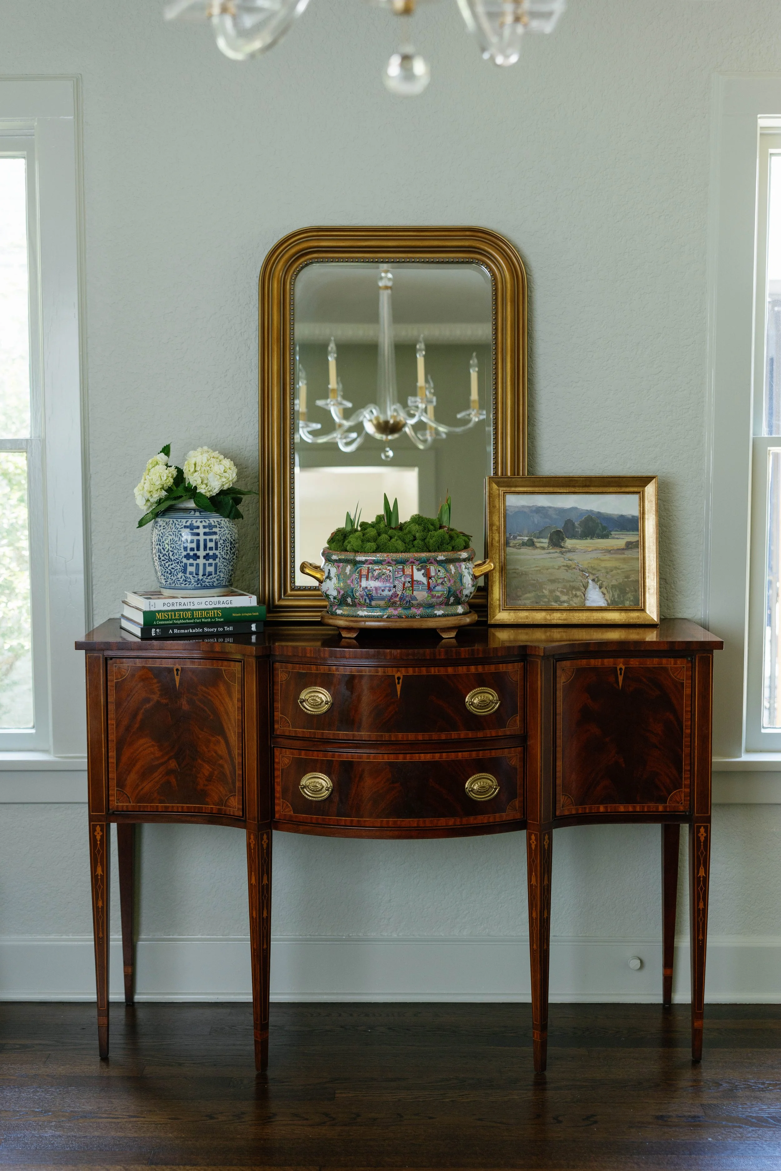 A wooden sideboard with a marble top, featuring a large gold-framed mirror hanging above it. The sideboard has three doors with ornate brass handles and intricate inlay, standing on four tapered legs. On the sideboard, there is a blue and white patterned vase with white hydrangeas, a stack of books, a decorative bowl with greenery, and a framed landscape painting. A chandelier is partially visible in the reflection of the mirror, and two windows flank the scene, letting in natural light.