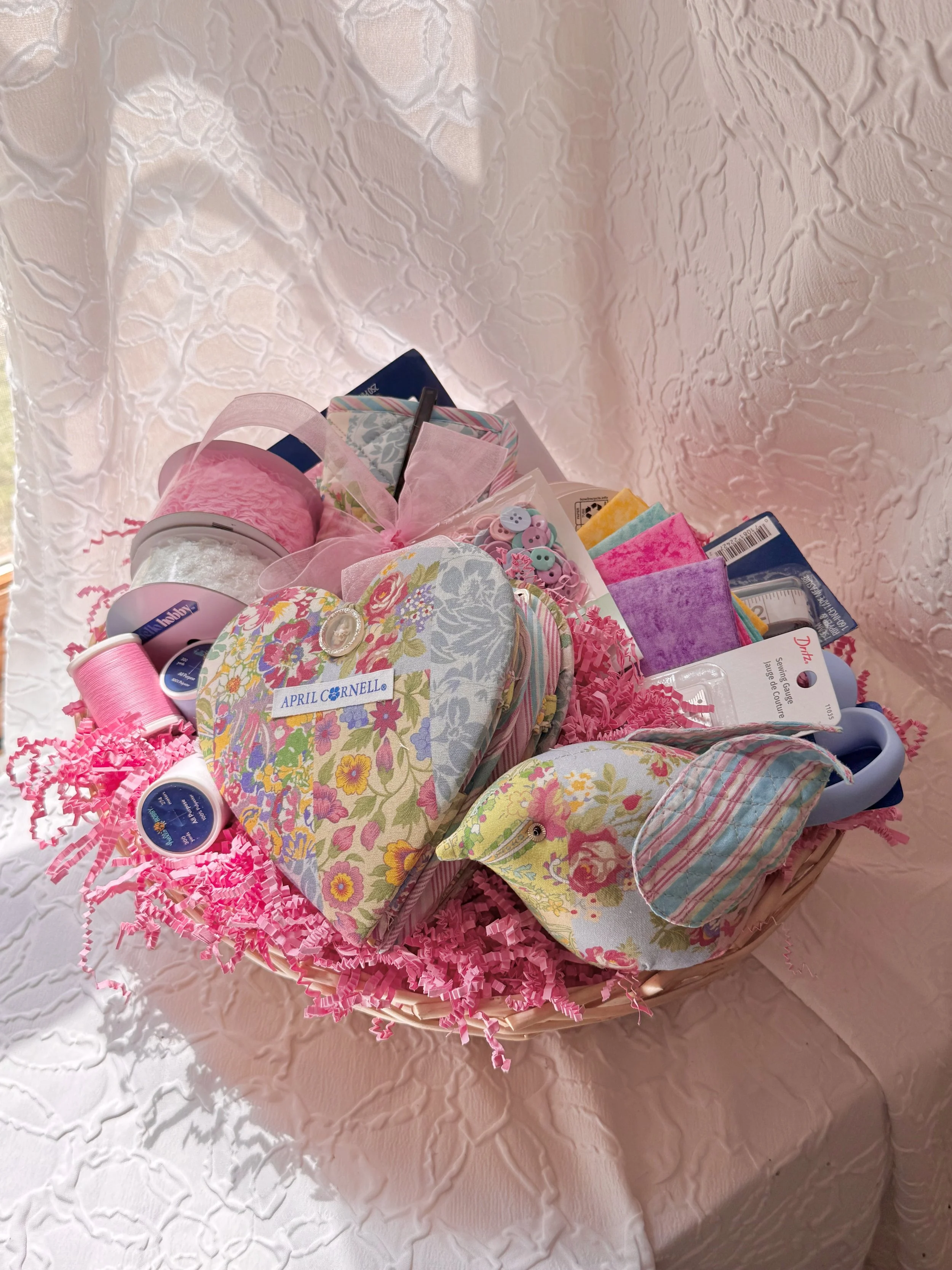 A basket filled with various sewing and craft supplies, including fabric, ribbons, buttons, and tools, sitting on a pink textured tablecloth.
