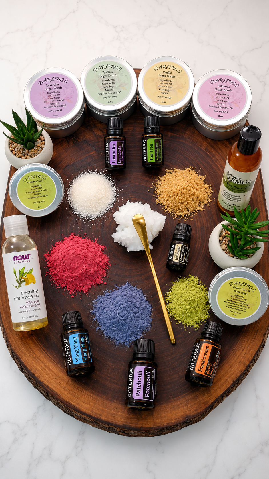 Arrangement of essential oils, colorful powders, and skincare ingredients on a circular wooden tray, with potted succulents and a gold spoon, on a marble surface.