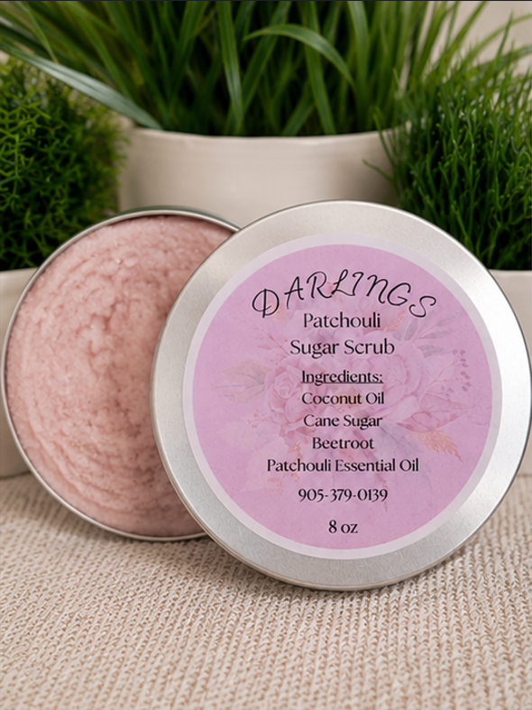 Open tin of patchouli sugar scrub labeled with ingredients: coconut oil, cane sugar, beetroot, patchouli essential oil, on a beige textured surface with green plants in the background.