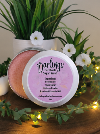Open tin of patchouli sugar scrub labeled with ingredients: coconut oil, cane sugar, beetroot, patchouli essential oil, on a beige textured surface with green plants in the background.