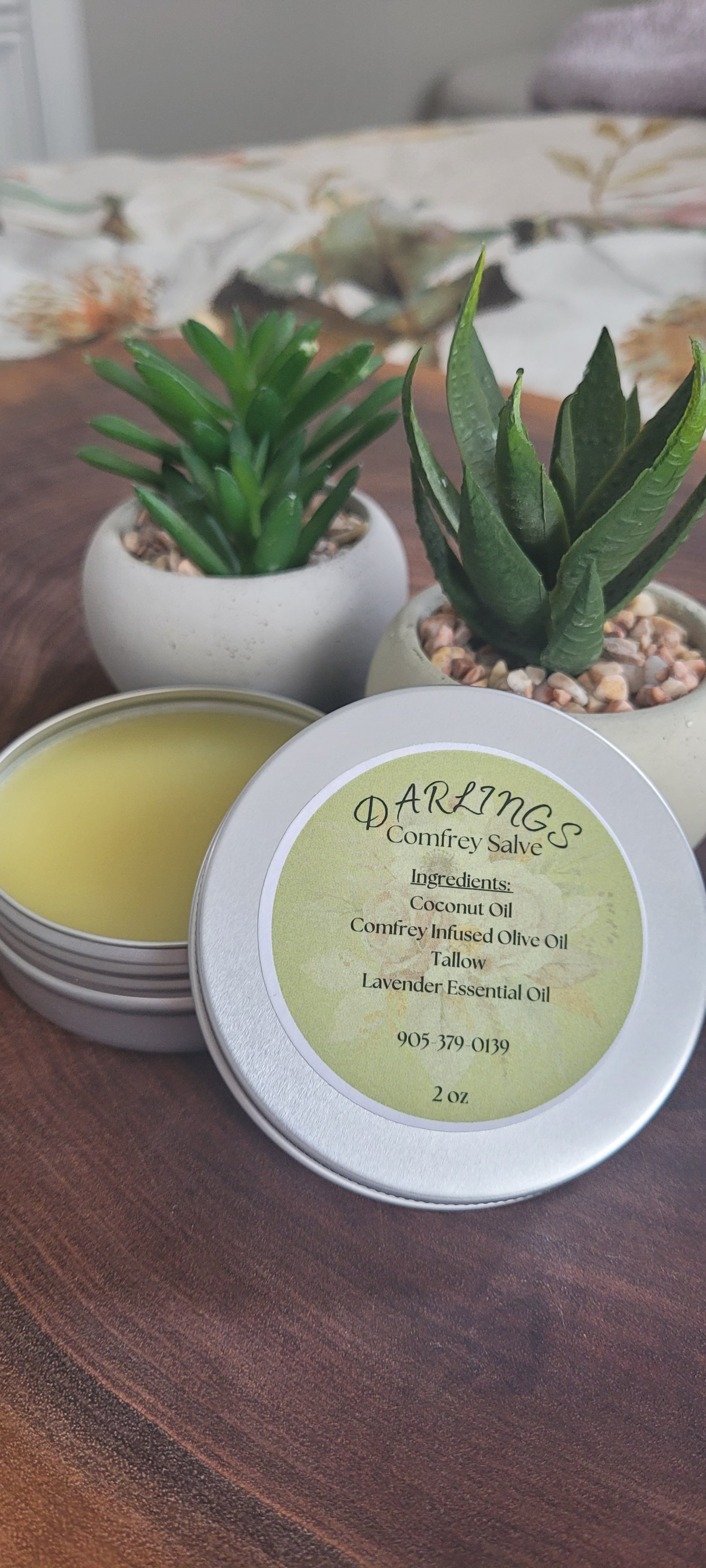 A tin of Darling's Comfrey Salve with ingredients listed on the lid, accompanied by two potted succulent plants on a wooden surface.