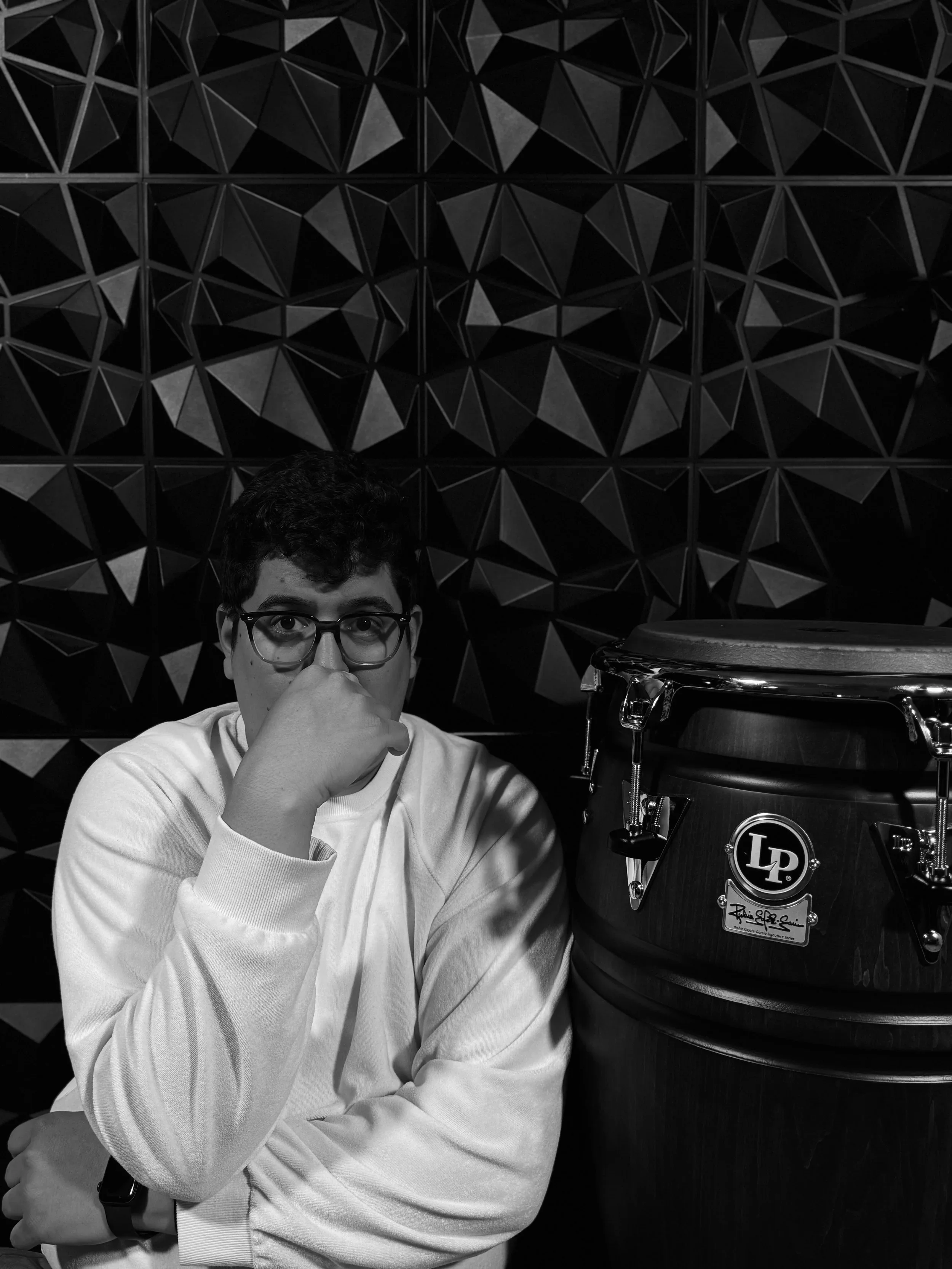 A young man wearing glasses and a light-colored sweatshirt sitting at a table with a percussion instrument, a large drum, next to him. The background features a geometric textured wall.