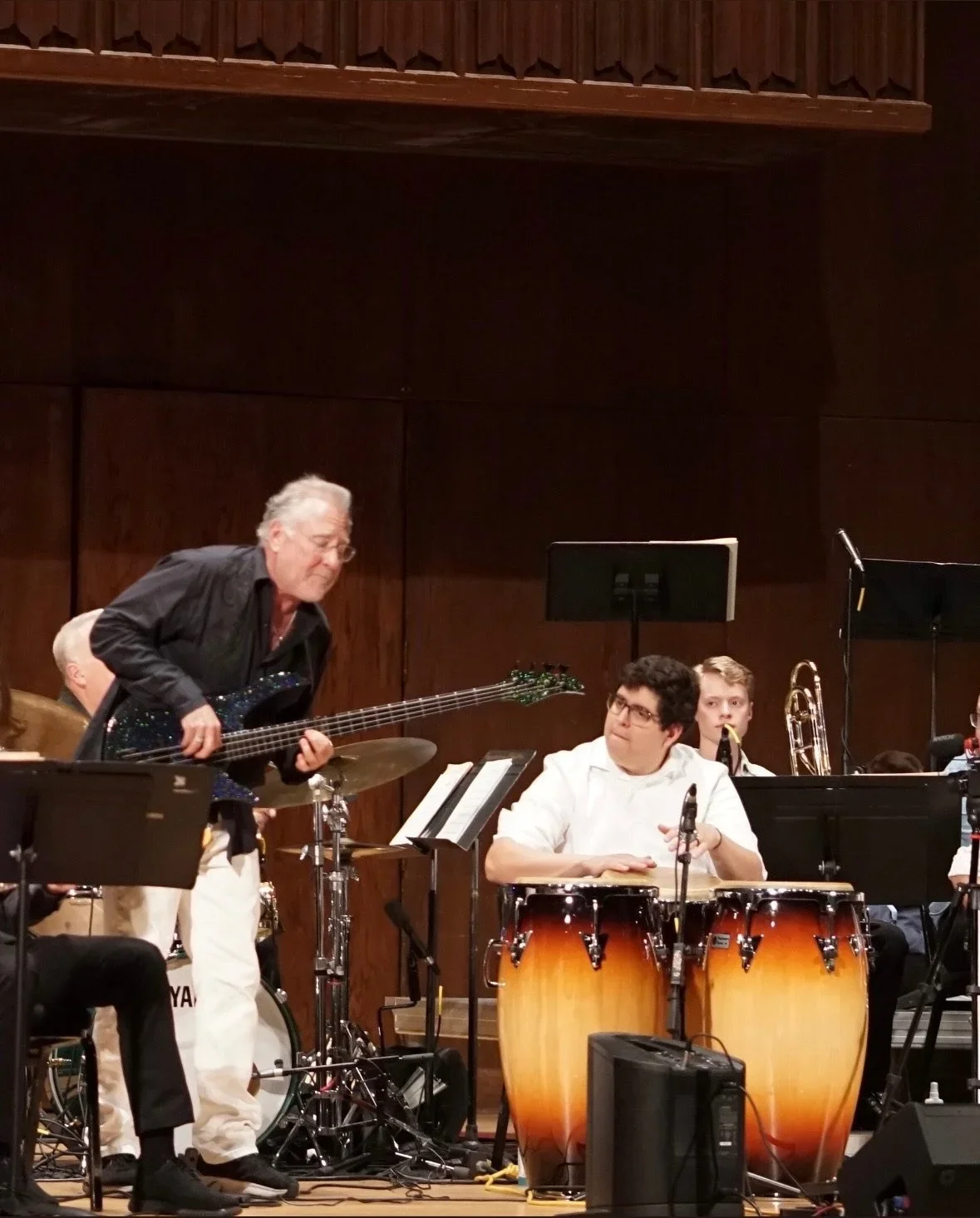 An older man is playing an electric bass guitar while a group of young musicians, including a percussionist, are seated behind him on a stage in a wood-paneled music hall.