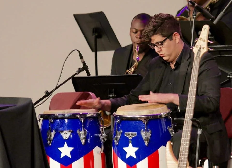 Two musicians playing percussion and wind instruments during a performance. The percussionist is playing bongos decorated with an American flag motif, and the other musician is playing a saxophone.