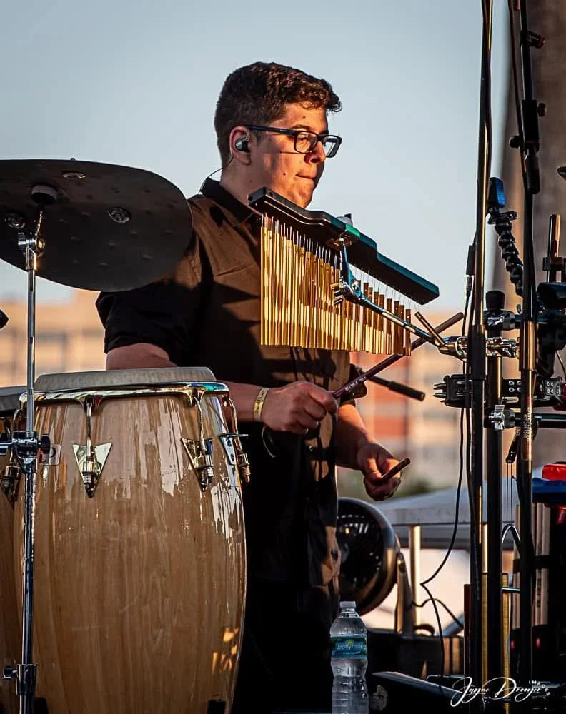 A young man playing a vibraphone, with a conga drum and a water bottle nearby, outdoors during sunset.