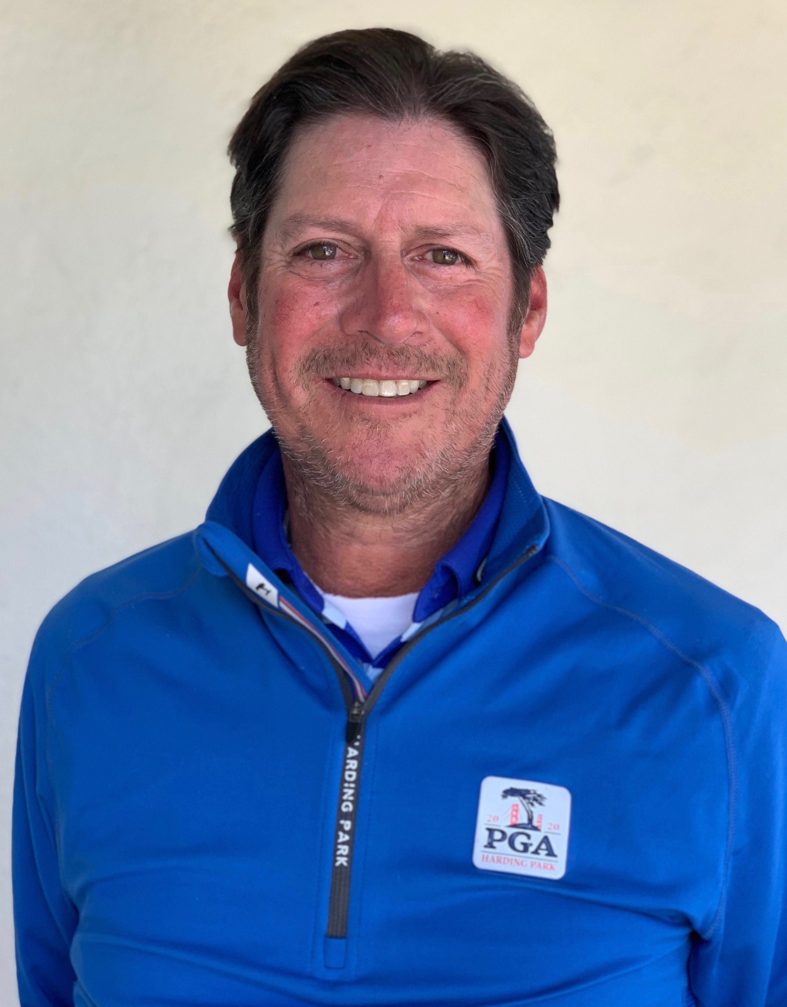 Close-up of a middle-aged man with dark hair and light brown eyes, smiling, wearing a blue jacket with a PGA logo and a badge.