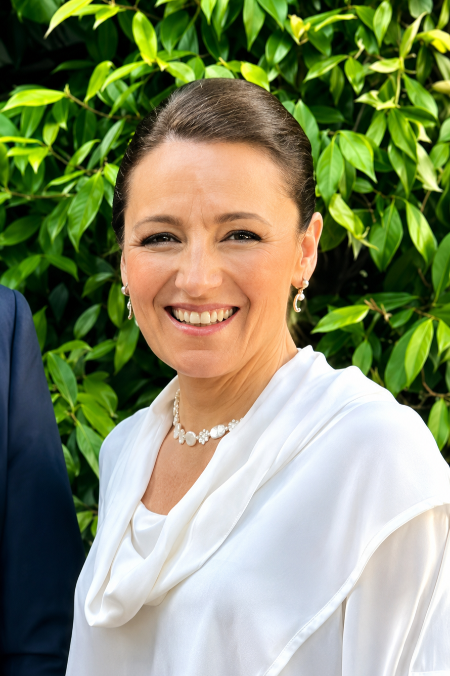 A smiling woman with dark hair tied back, wearing pearl jewelry, a white satin blouse with a draped neckline, standing outdoors in front of green leaves.