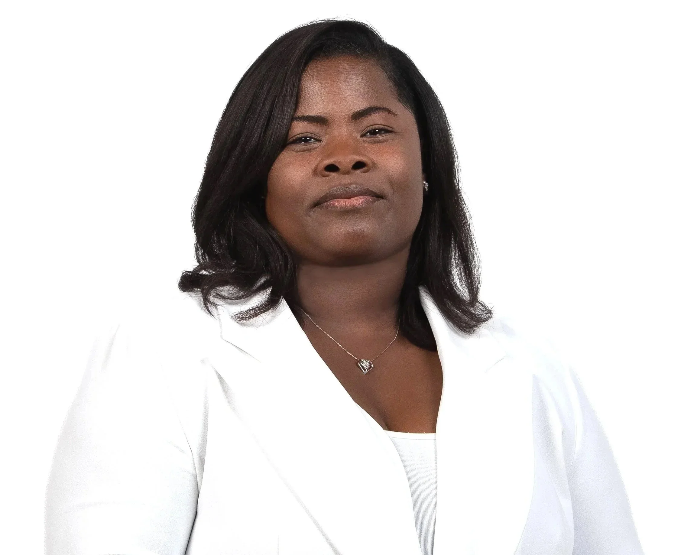 A woman with dark hair and dark skin wearing a white blazer, a white top, and a silver heart necklace, posing against a plain white background.