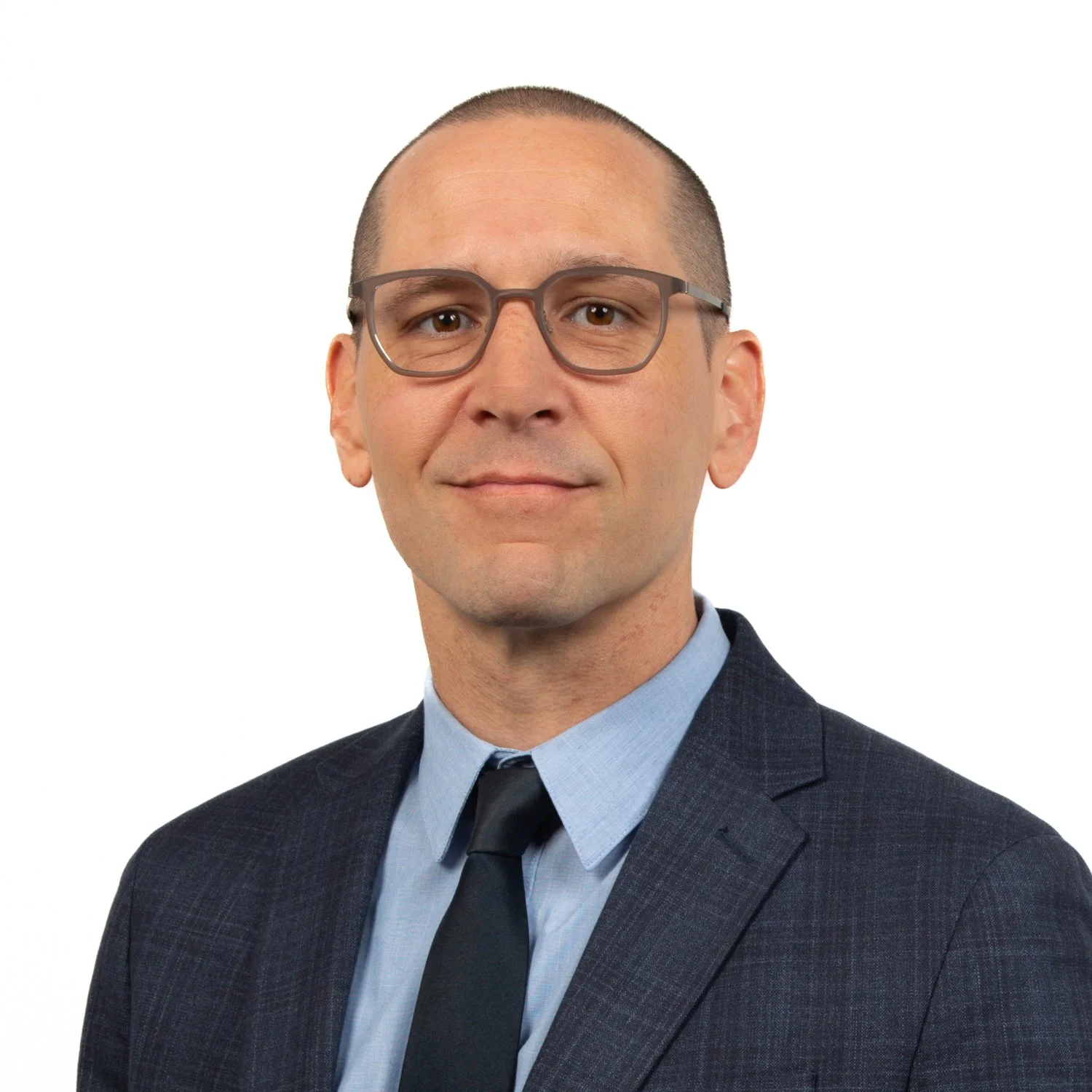 A man with glasses wearing a dark suit, light blue shirt, and dark tie posing against a white background.