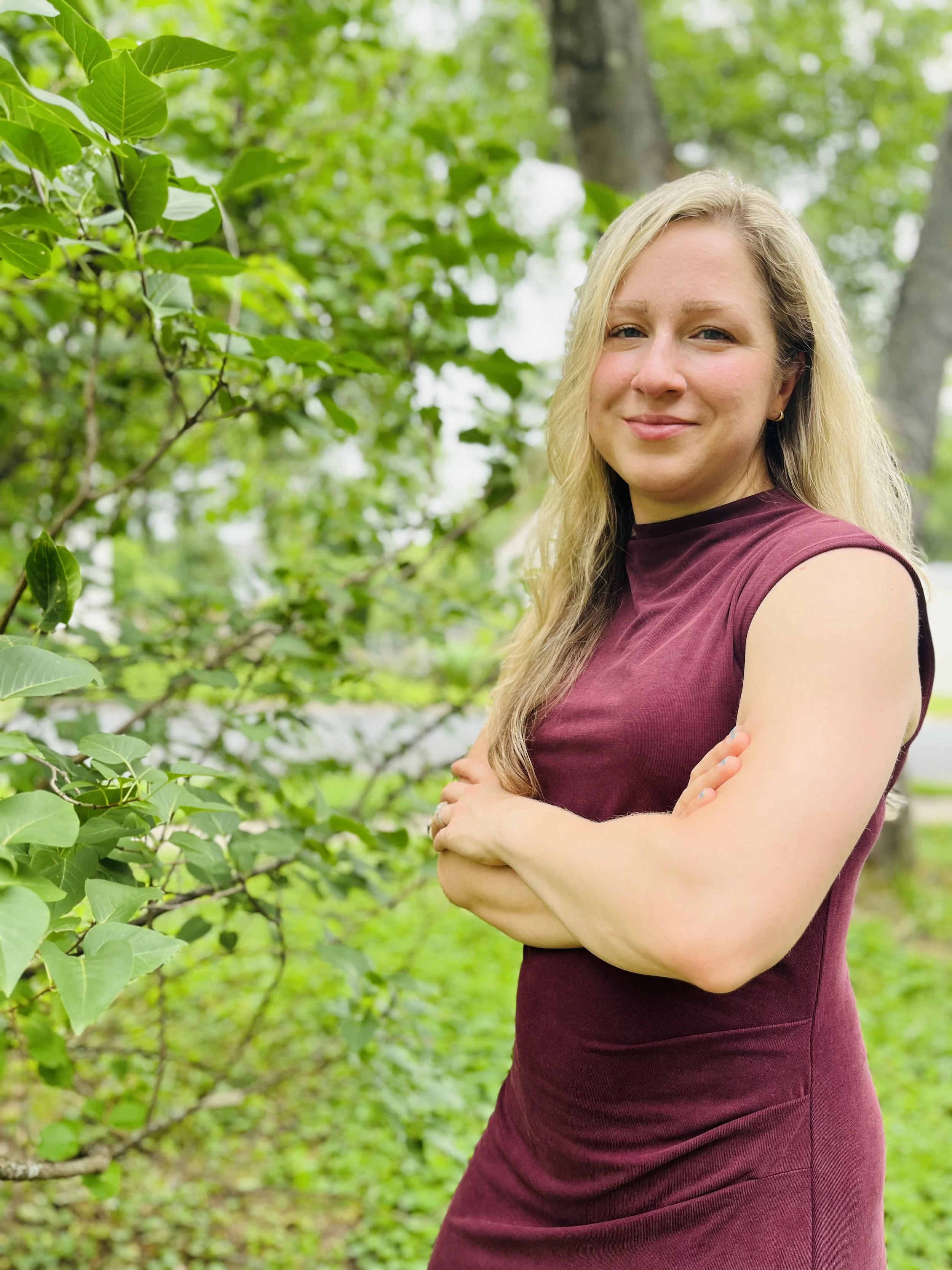 A blonde woman wearing a sleeveless maroon dress standing outdoors among green trees with her arms crossed and smiling.