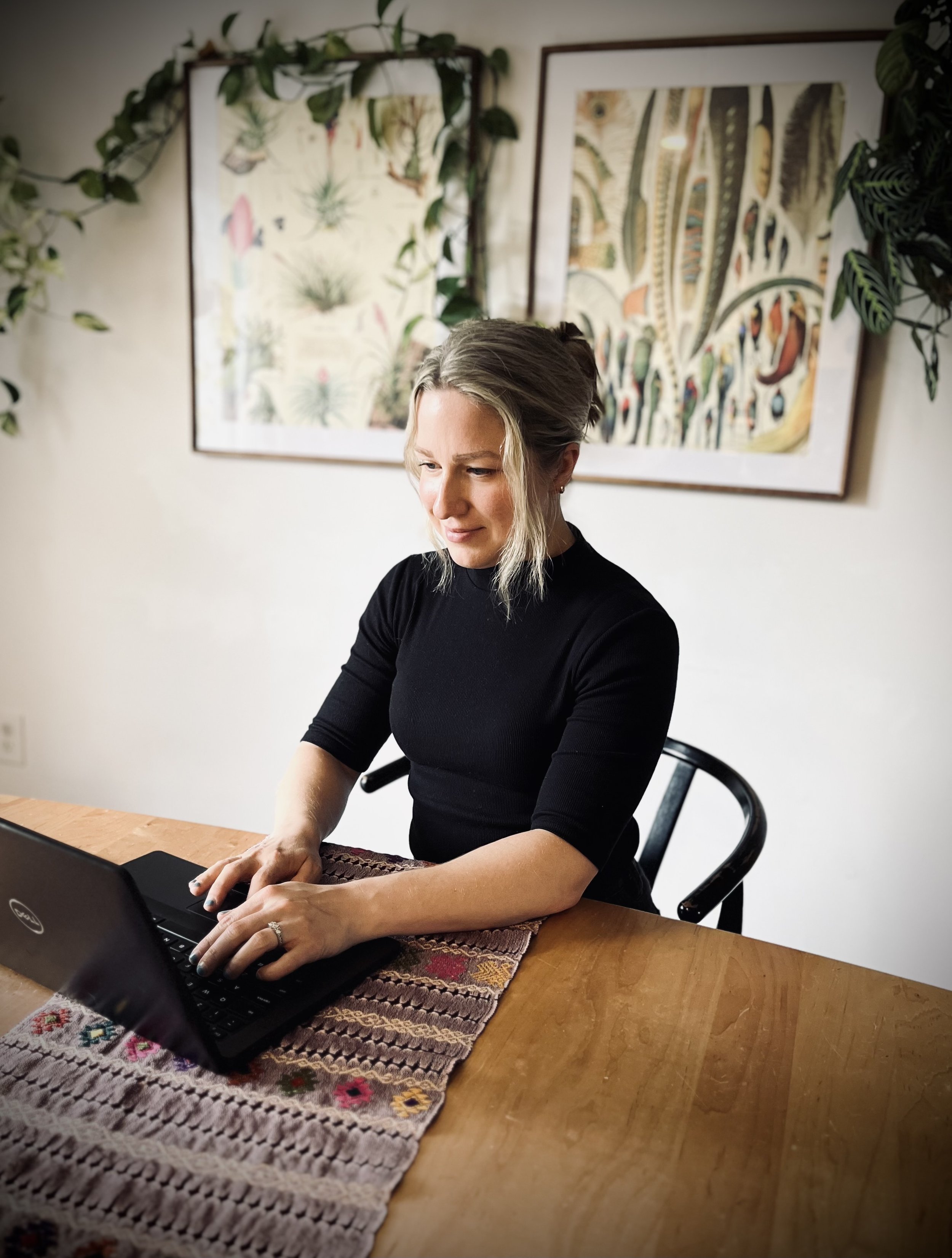 A woman with blonde hair working on a laptop at a wooden table, with a colorful, patterned tablecloth. Behind her, there are two framed botanical art prints on a white wall and some green leafy plants.