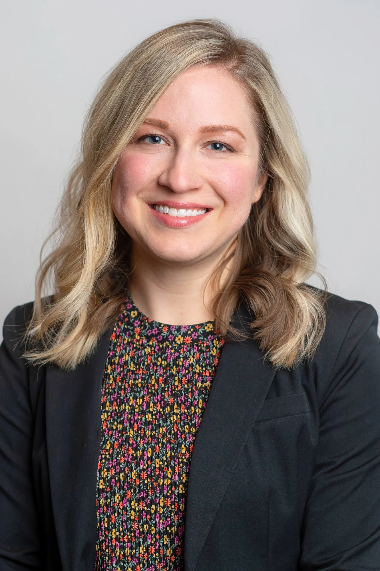 Close-up portrait of a woman with blonde hair and blue eyes, smiling, wearing a black blazer and a floral-patterned blouse, against a plain light gray background.