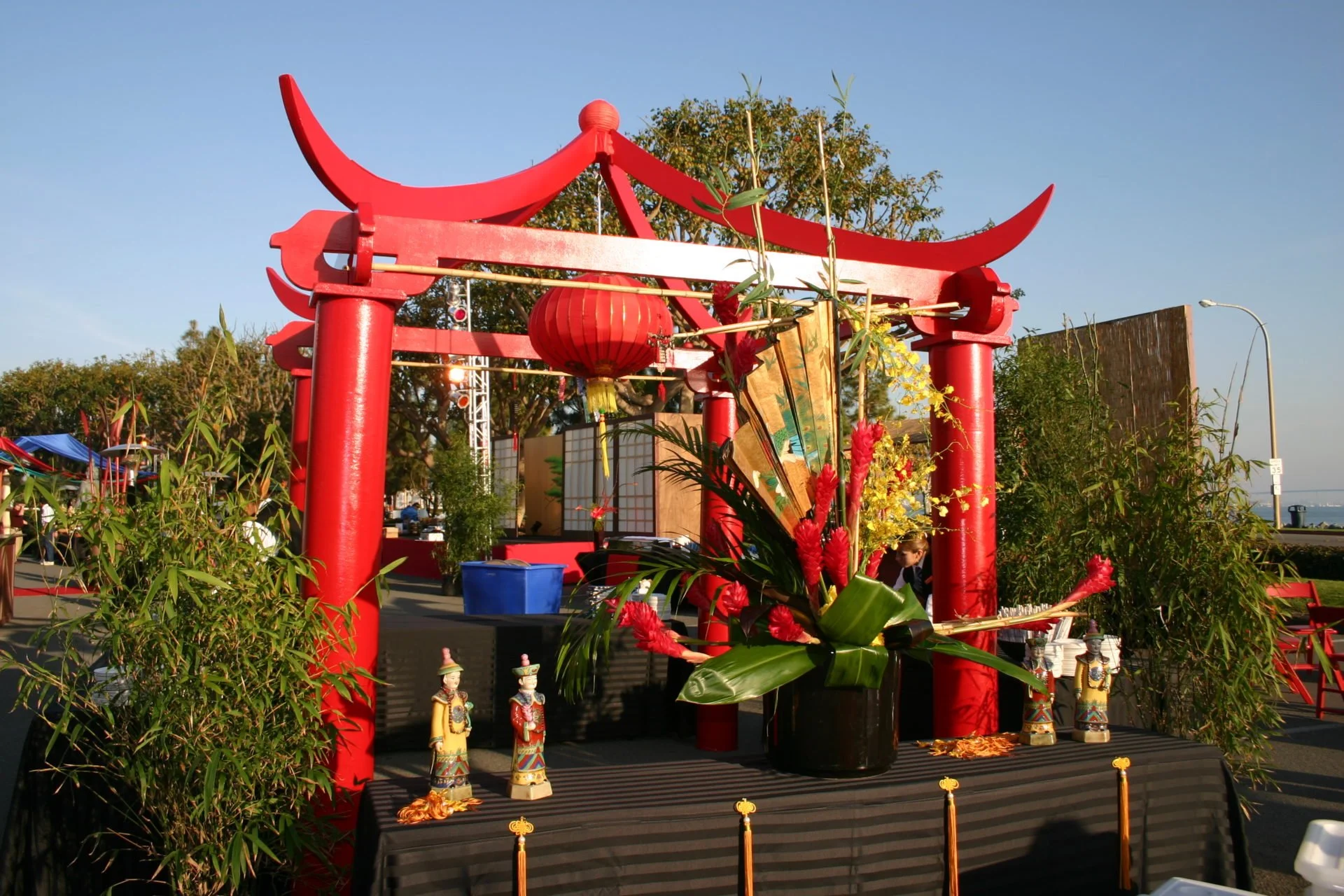 Red Chinese archway with decorations including a red lantern, flowers, and figurines, outdoor setting with trees and a clear sky.