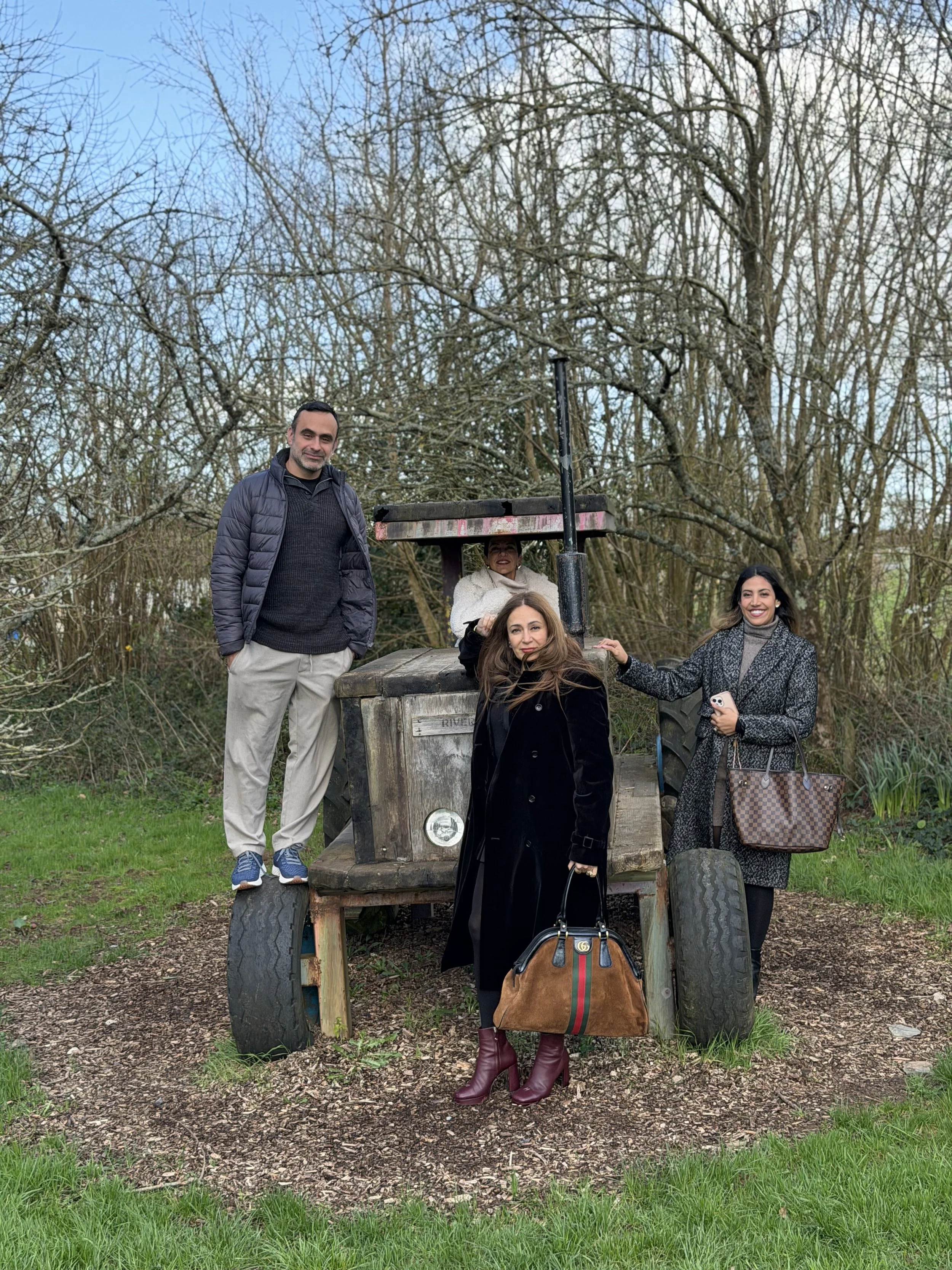 Four people posing outdoors in a park with leafless trees in the background. They are standing around an old, rustic vehicle or structure, with one person sitting or reclining on it. The individuals are dressed in casual and stylish clothes, some hol