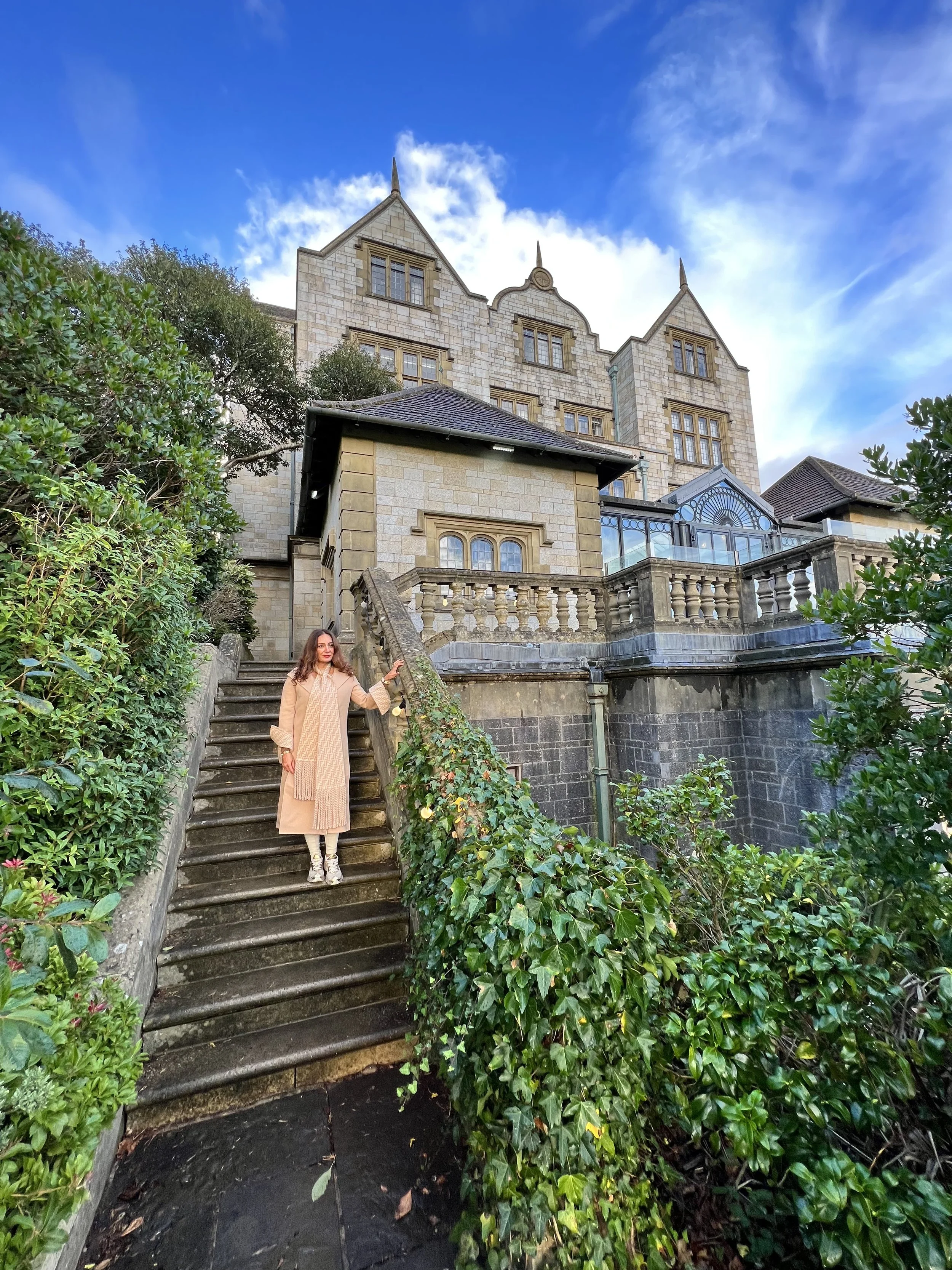 A woman with long brown hair wearing a beige coat and sneakers stands on stone stairs with ivy on both sides, leading up to a large historic stone mansion with multiple windows and pointed rooftops, under a partly cloudy blue sky.